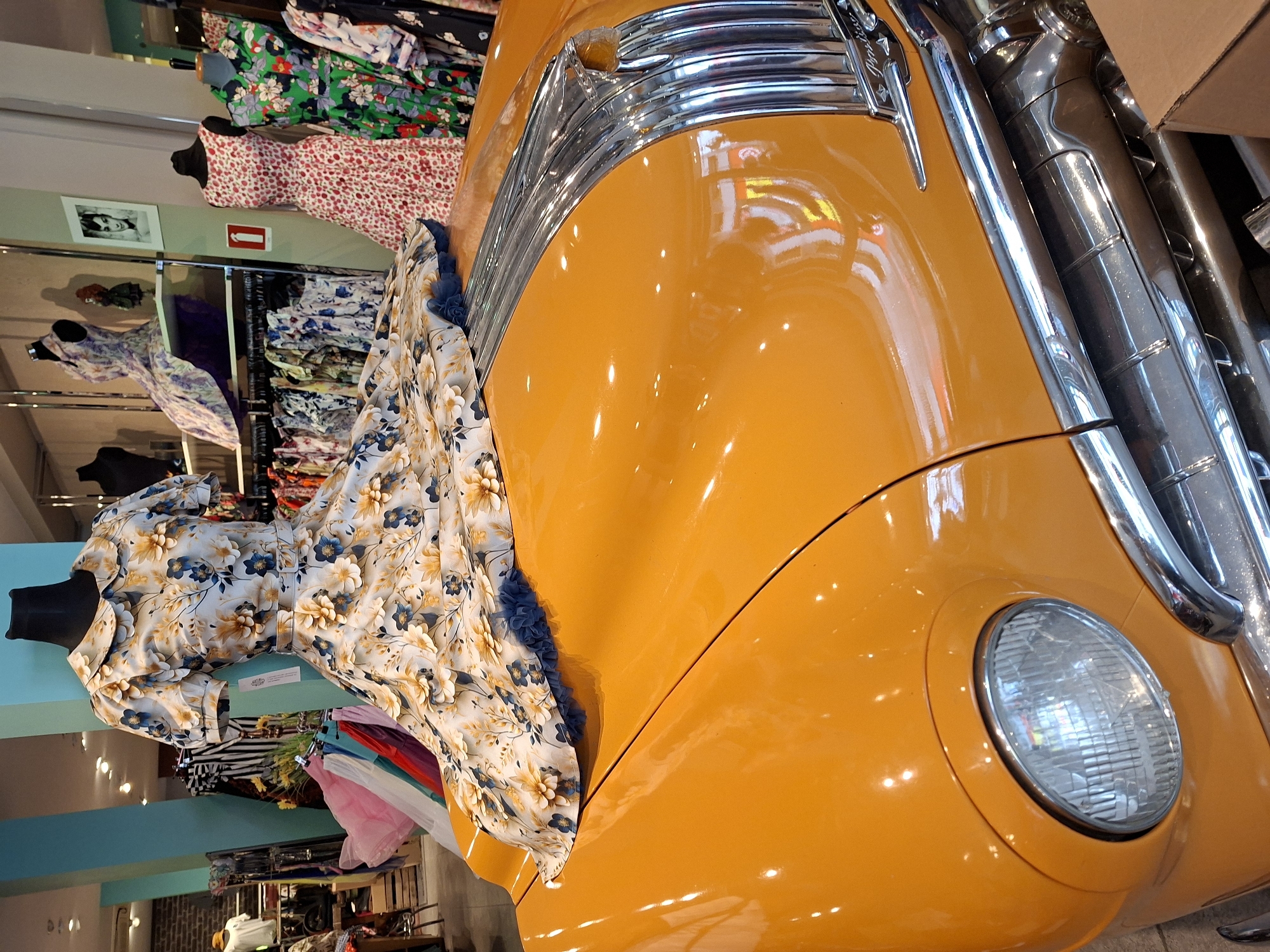 The image shows a vintage orange car with a floral print dress displayed on its hood. The car's hood is highly reflective, showing distortions of the indoor environment. The dress is light-colored with a pattern of yellow and blue flowers and has blue ruffled trim along the hem that drapes over the car. In the background, mannequins are visible wearing dresses of various styles and colors, suggesting a retail store. Shelves with more clothing and a framed picture of Audrey Hepburn are also present. The store appears to be a boutique. This scene is located at the vintage clothing store "The Old Town" in Ostend, Belgium.