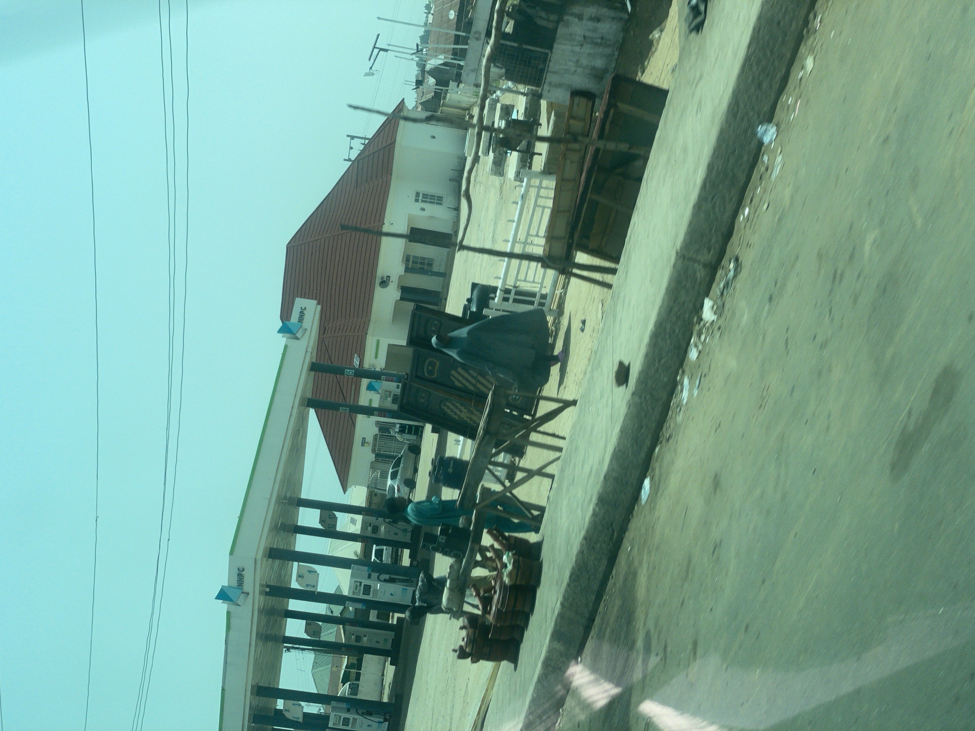 The image captures a scene at an NNPC gas station in Bauchi, Nigeria. In the foreground, a person in a dark blue, flowing garment walks away from the camera, past a display of what appear to be wooden barrels and other items arranged on the ground and on a makeshift wooden stand. To the left, another person in a teal-colored garment stands near the barrels. In the background, the gas station canopy with "NNPC" signage is visible, along with fuel pumps. Beyond the gas station, a white building with a reddish-brown tiled roof is present. The overall environment appears to be an outdoor urban setting with a slightly hazy or filtered sky, suggesting either a bright day or a reflection through glass.