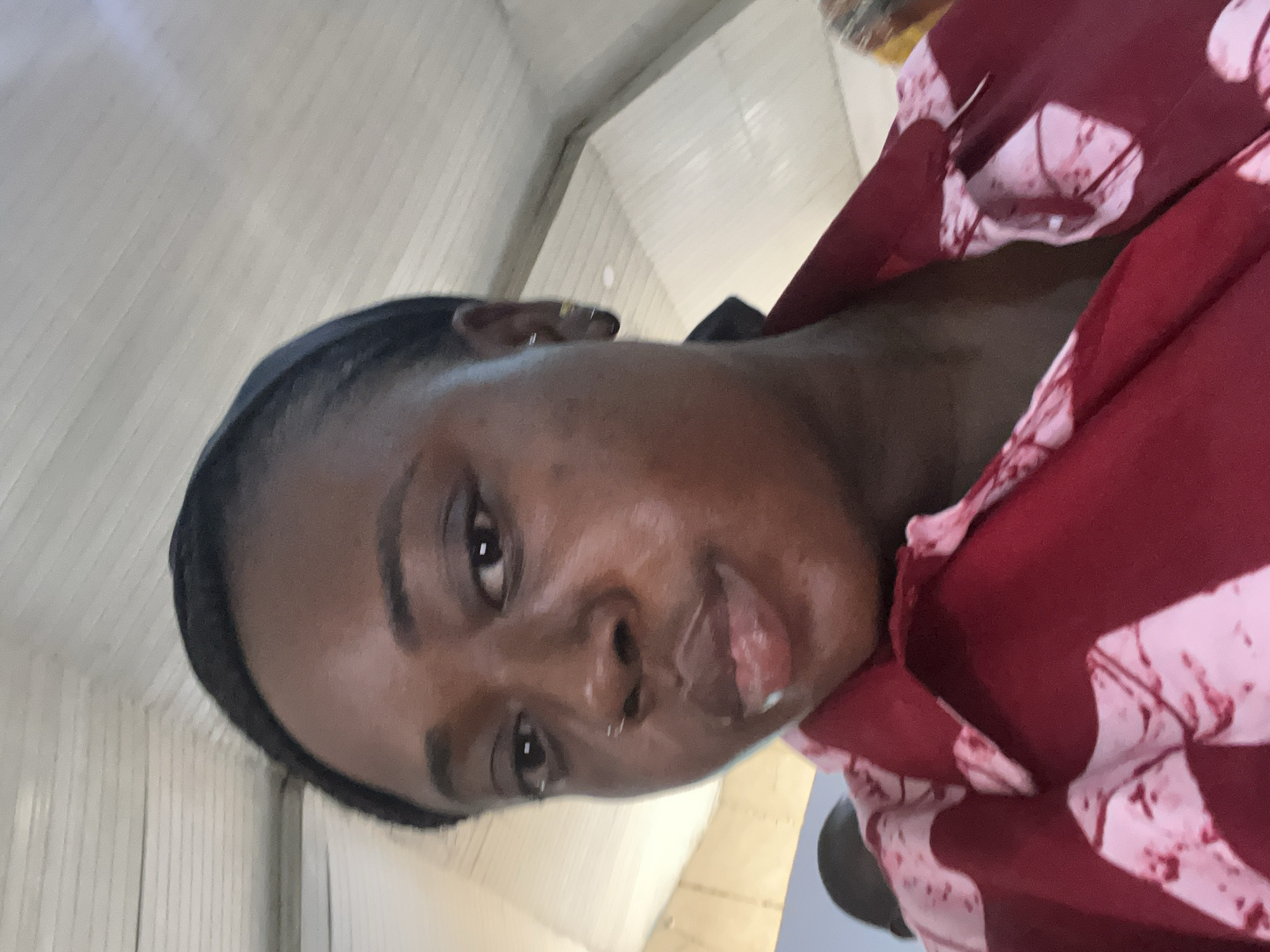 A close-up, slightly angled portrait shows a dark-skinned woman with short, dark hair and a nose ring. She is wearing a dark headband and a patterned garment with red and white colors. The background appears to be an interior space with light-colored walls, potentially a building. There is no verifiable information about the location being in Jos, Nigeria or anything else.
