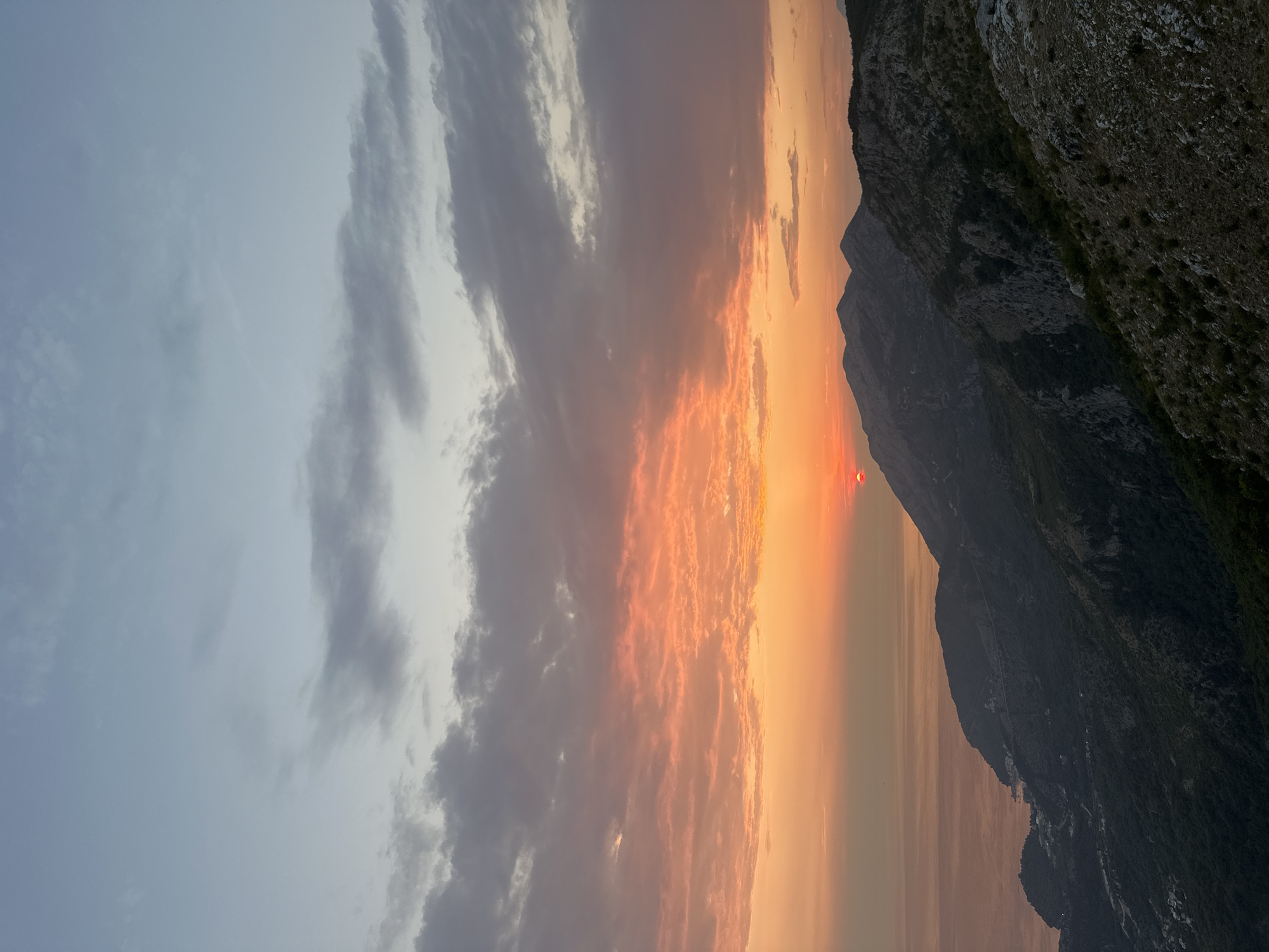 This image captures a panoramic view of a mountainous coastal landscape at sunset, likely near Đurovići, Montenegro. The sky is filled with dramatic clouds painted in hues of orange, pink, and gray, transitioning to a pale blue at the zenith. A sliver of the setting sun, a vibrant red orb, is visible just above the horizon line, casting a warm glow over the scene.
Below the sky, a rugged mountain range dominates the mid-ground and foreground. The mountains are characterized by steep slopes, rocky outcrops, and patches of dark green vegetation. The texture of the terrain suggests a wild, untamed environment. In the distance, a body of water, likely the Adriatic Sea, is visible, reflecting the soft colors of the sunset. Along the coastline and nestled within the foothills of the mountains, faint signs of human habitation can be discerned, appearing as small clusters of buildings or structures. The overall atmosphere is serene and breathtaking, highlighting the natural beauty of the Montenegrin coast. There are no people or specific objects or actions depicted in the image.