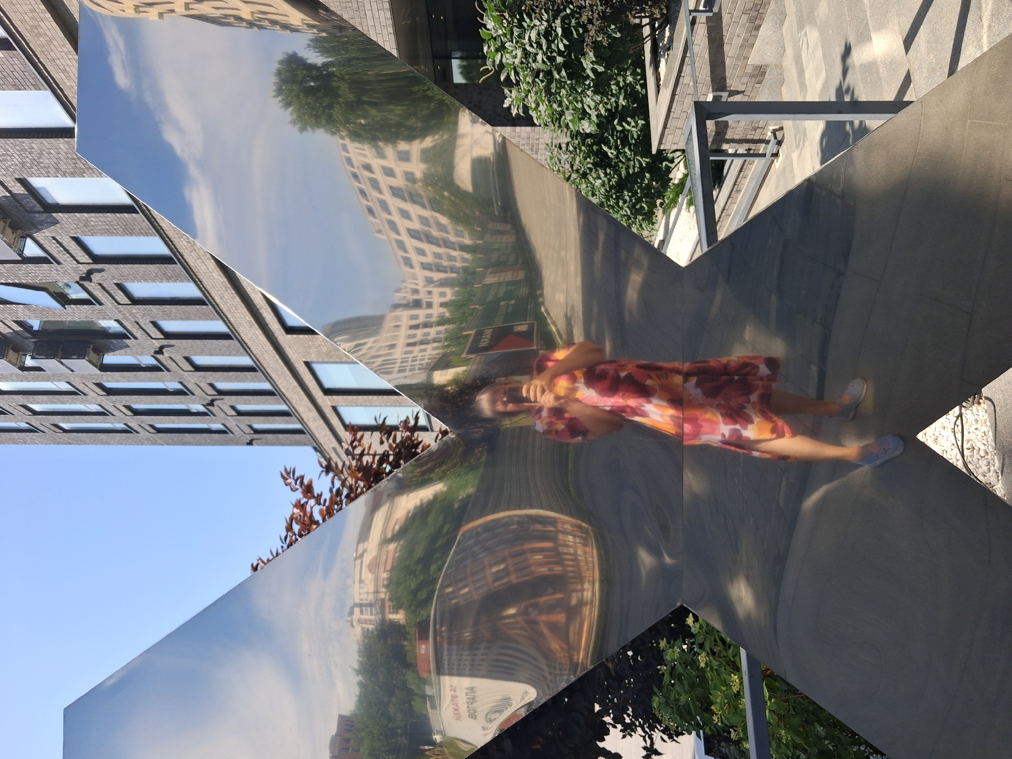 A woman in a floral dress is reflected in a large, angular, mirrored sculpture outdoors on a sunny day. The sculpture is composed of several flat, reflective surfaces angled to create a fragmented view of the surroundings. The woman is holding a phone in her right hand, presumably to take the photo. She is standing on a paved area, with greenery and buildings visible in the reflections. The background shows a modern brick building and a clear blue sky with some clouds. The location is identified as Novosibirsk, Russia.