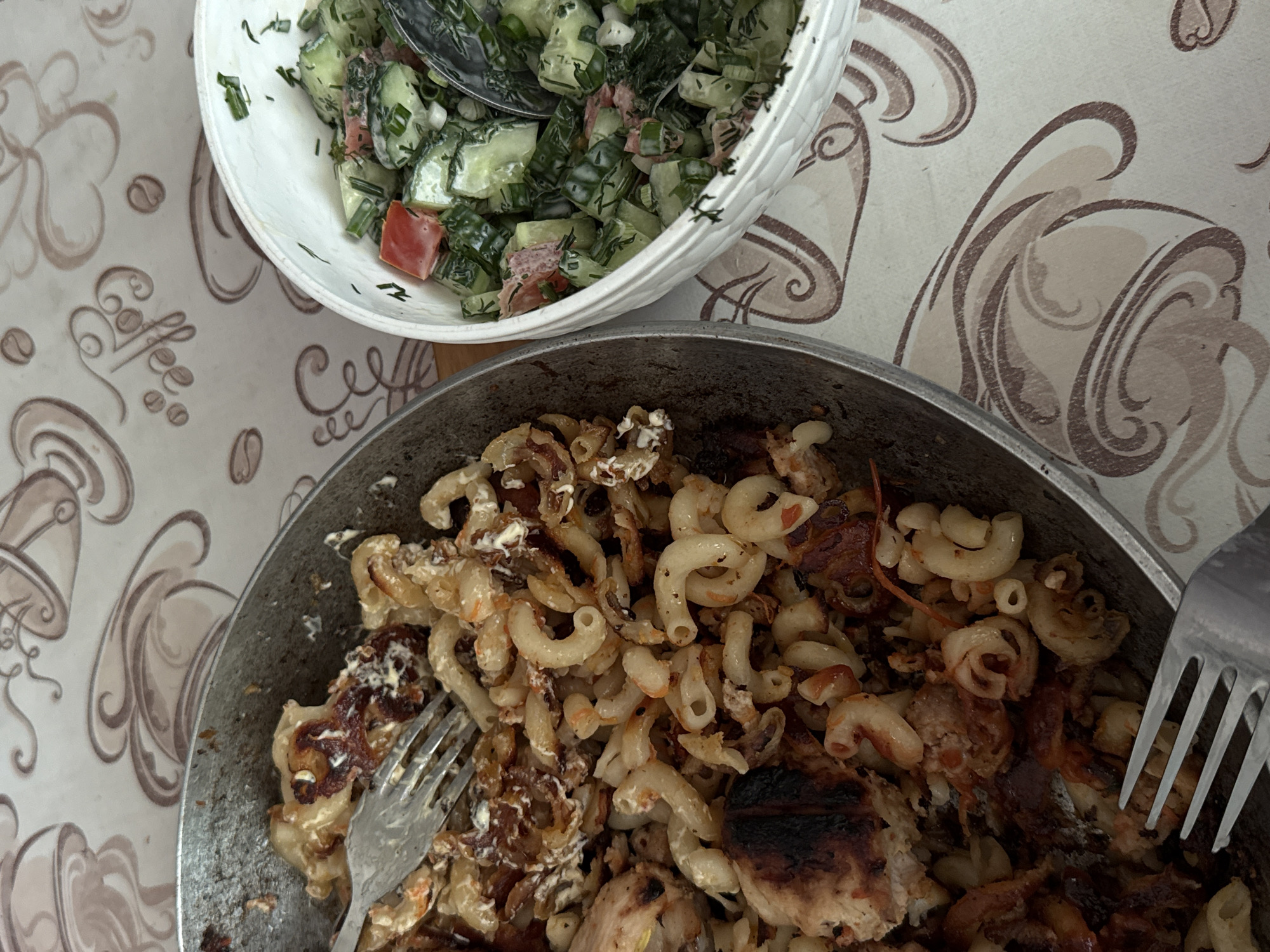 A top-down shot depicts a meal on a patterned tablecloth, possibly taken indoors. In the upper left, a white bowl contains a salad with cucumbers, tomatoes, and greens. Below, a frying pan holds cooked pasta, possibly elbow macaroni, with fried meat and onions. Two forks are visible, one embedded in the pasta and the other alongside the pan. The tablecloth displays a coffee bean and coffee cup design. The location is likely a private residence, not specifically identifiable as Uvarovo, Russia without additional context.