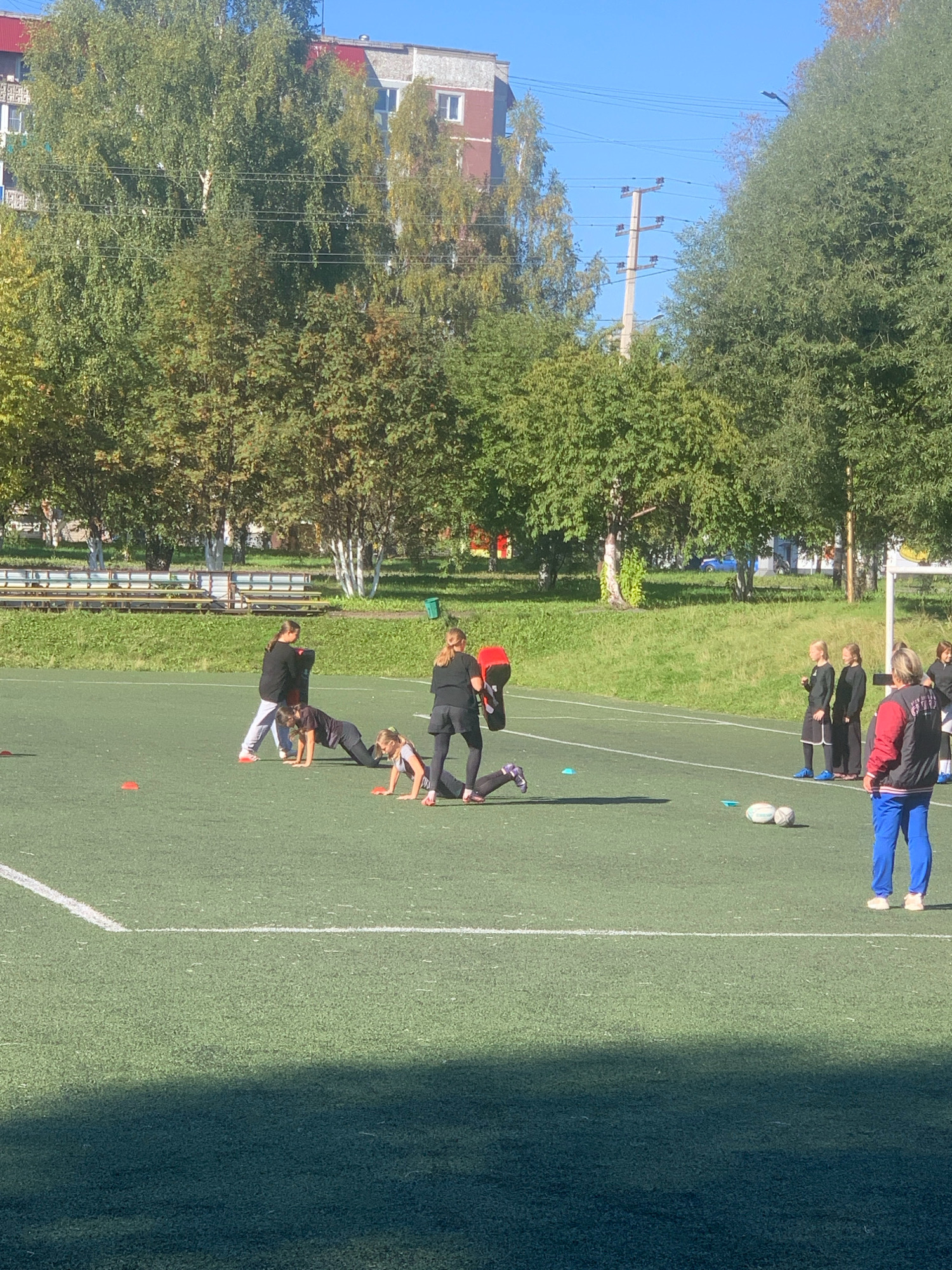 The image shows an outdoor sports field with artificial turf and white markings. Several young people, appearing to be boys and girls, are engaged in physical training. One individual is holding a red kicking pad, suggesting a martial arts or combat training session. In the background, there are trees, residential buildings with red roofs, and electrical poles. The scene is illuminated by natural sunlight, casting shadows on the field. This outdoor sports facility is likely located in the city of Prokopyevsk, Russia, given the context provided.