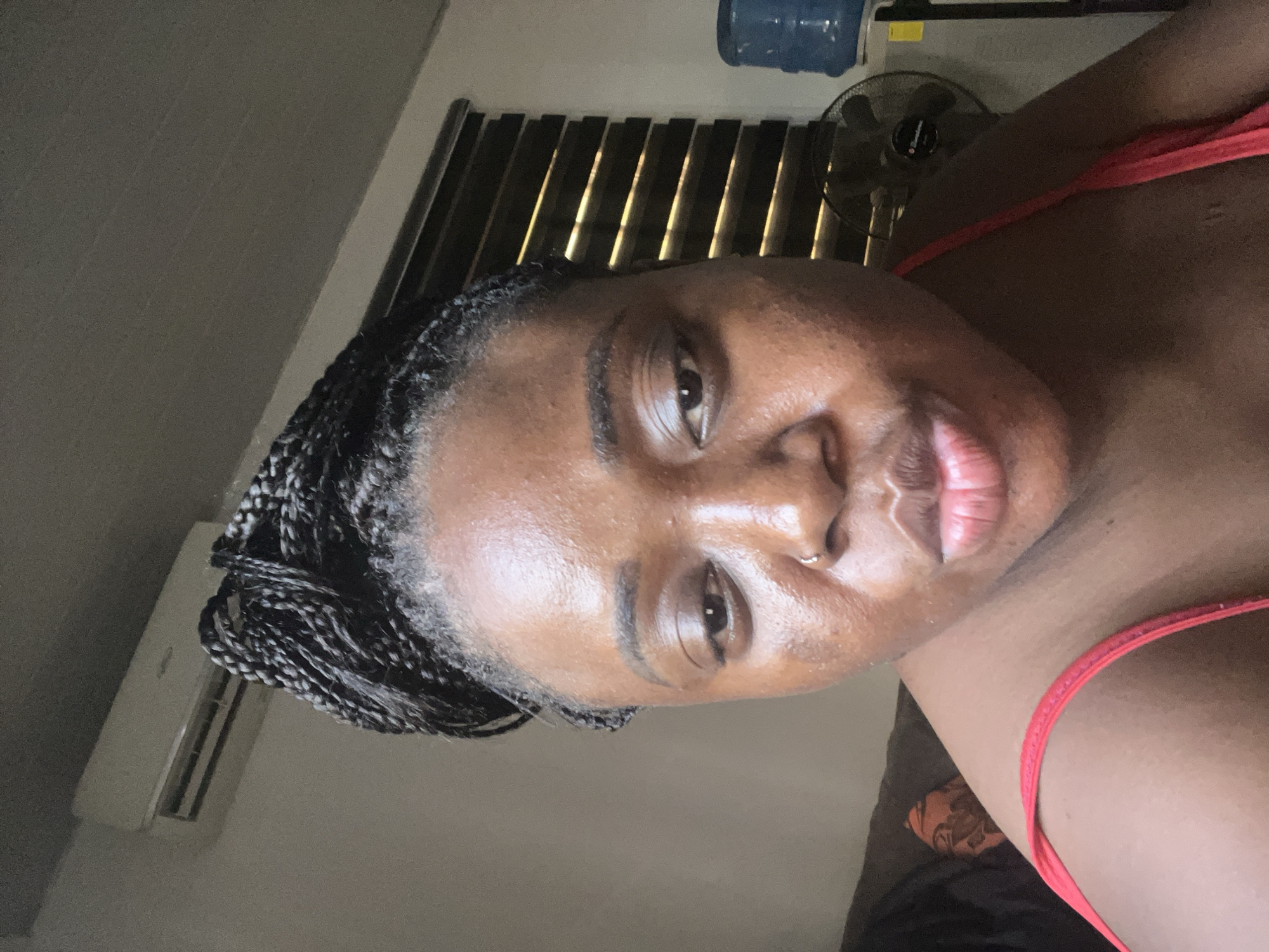 The image shows a close-up of a person with dark skin, braided hair, and a nose ring, wearing a red top. The background includes a window with closed blinds, a fan, and a water dispenser. An air conditioner is visible on the wall in the lower-left corner. Without additional details, it's impossible to pinpoint the specific location within Abuja, Nigeria.
