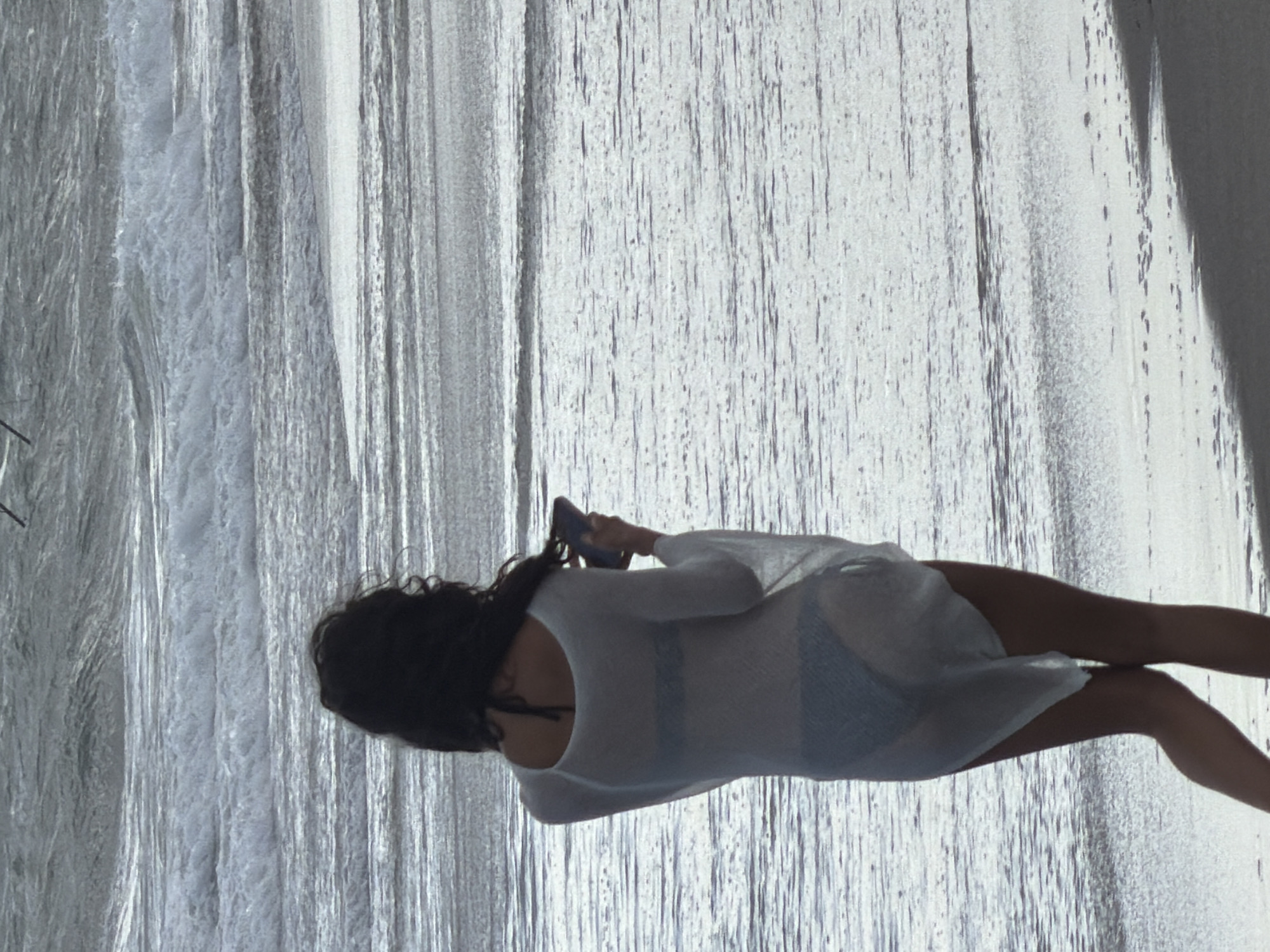 A woman stands on a wet sandy beach facing away from the camera, looking out at the ocean. She has long dark hair and is wearing a white sheer cover-up over a dark bikini. In her right hand, she holds a dark-colored smartphone. The ocean behind her is choppy with white-capped waves. The sky is overcast and appears to be gray. The sand in the foreground is dark and wet, indicating the tide is out or has recently receded. The image appears to be taken during the day, likely in the afternoon given the light. The location is the beach in North Kuta, Indonesia.