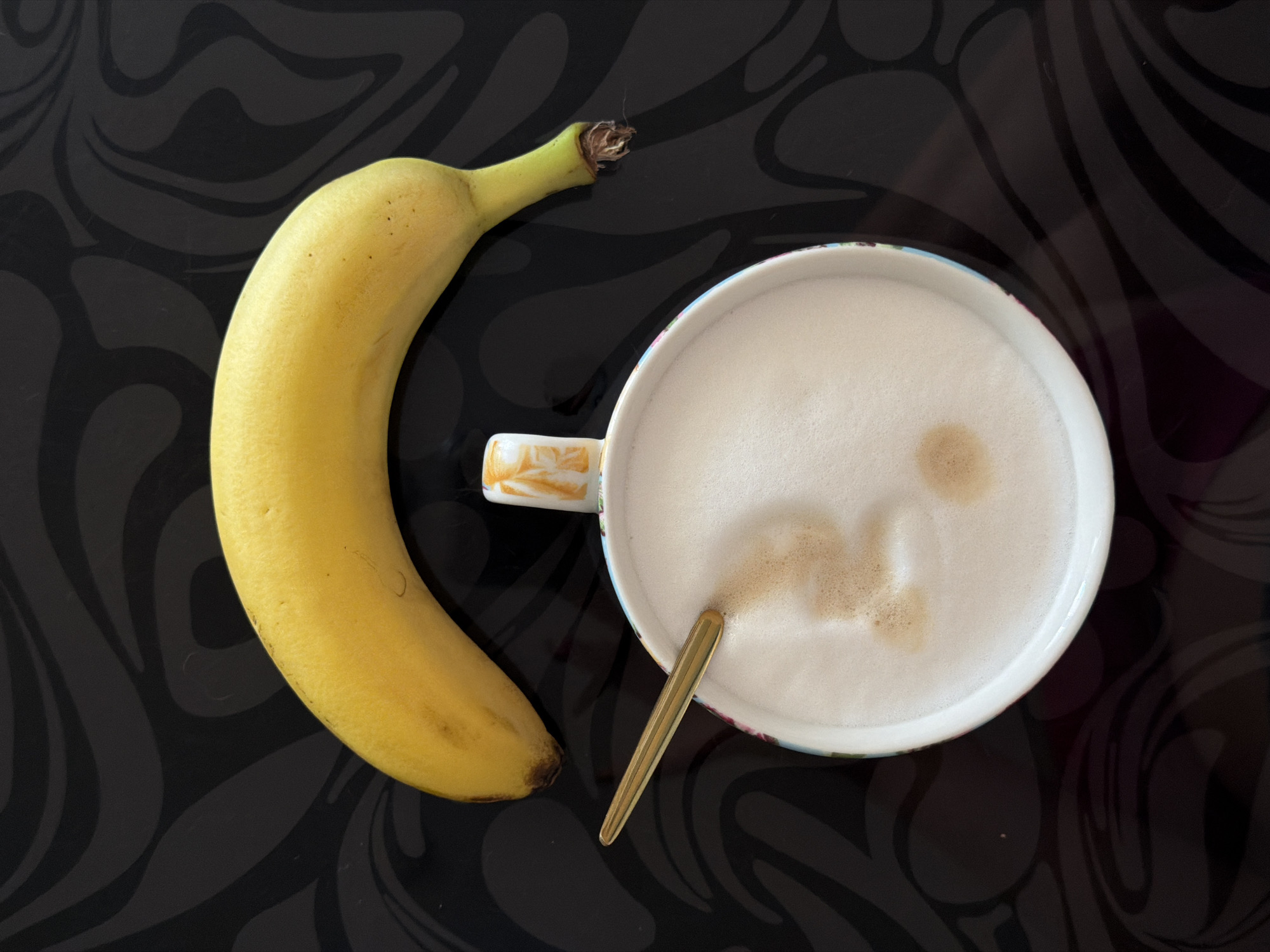 A banana and a cup of coffee with a spoon are placed on a patterned black surface. The banana is yellow and positioned to the left of the cup. The coffee appears to be a latte, with a frothy white surface and some brown coffee showing. A golden spoon is partially submerged in the coffee. The surface underneath has a dark, wavy pattern. The location of Rossiyskiy, Russia is not discernible from the image.