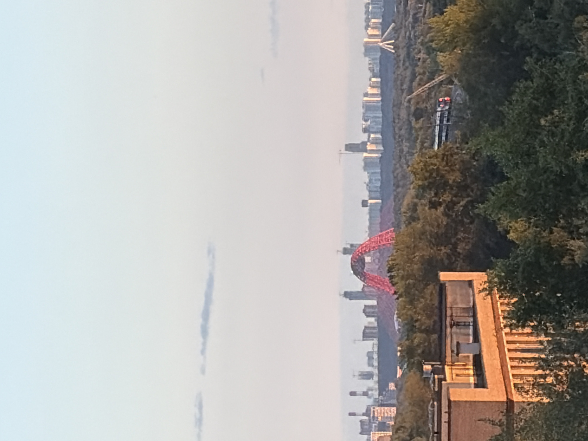 This image depicts a panoramic view of the city of Khimki, Russia, under a clear sky at sunset. In the foreground, a brick building with a flat roof is partially visible, surrounded by lush green trees. The middle ground showcases a dense forest with a train track cutting through it, on which a train with illuminated red lights is traveling. In the distance, the Khimki skyline is dominated by various skyscrapers and the iconic red arched bridge, also known as the Zhivopisny Bridge. The sky transitions from a pale blue at the top to a soft orange hue near the horizon, indicating the late hour of the day. The overall scene is serene and captures the urban landscape of Khimki bathed in the warm glow of the setting sun.