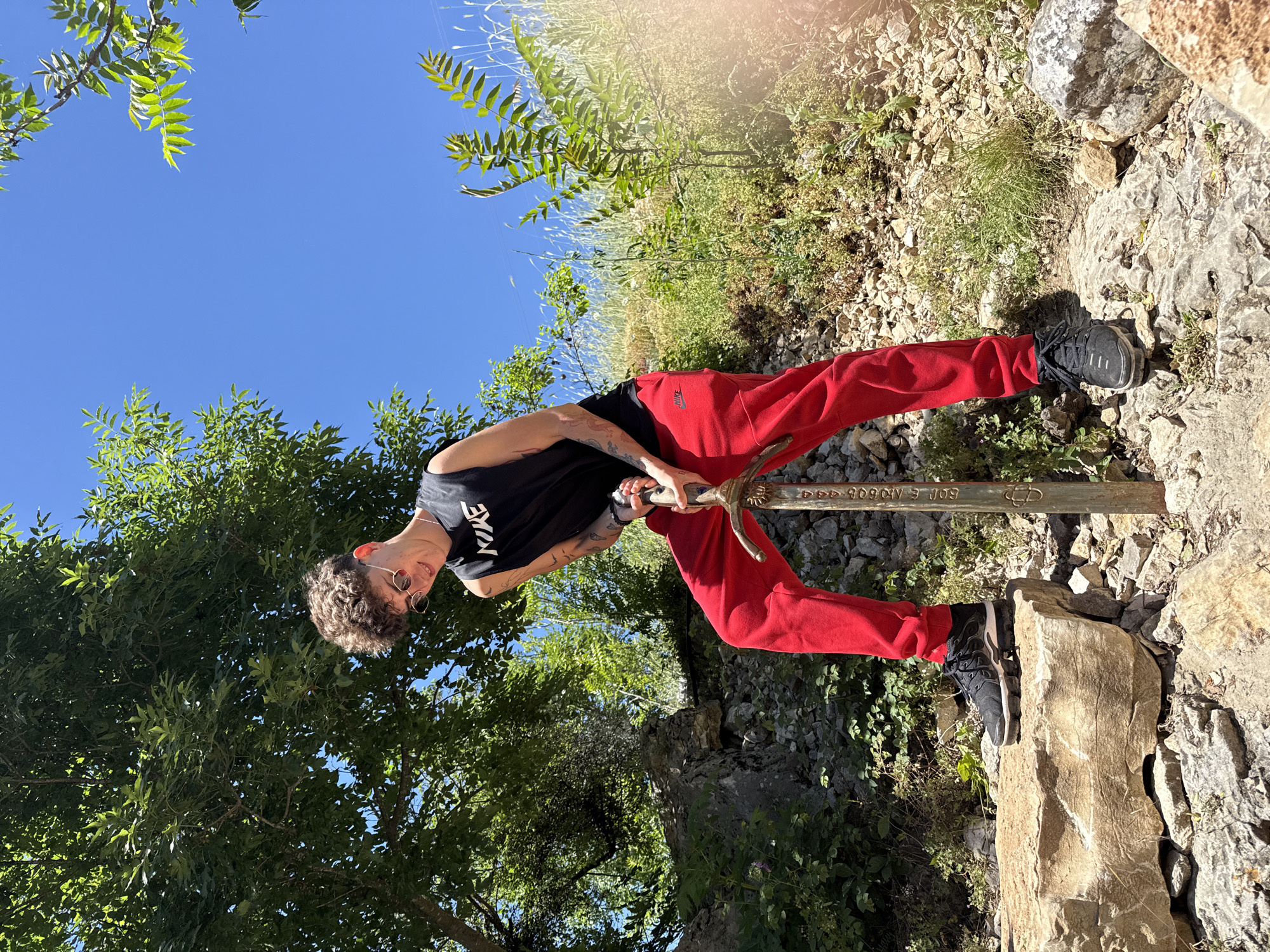 A young man stands with a sword, appearing to pull it from a rock. He is wearing a black tank top with the word "NIKE" visible, red sweatpants, and black sneakers. He also wears sunglasses and has short, curly hair. He is situated on a rocky terrain covered with grass, stones, and some shrubs. The sky is clear and blue. The location is Lukovit, Bulgaria.