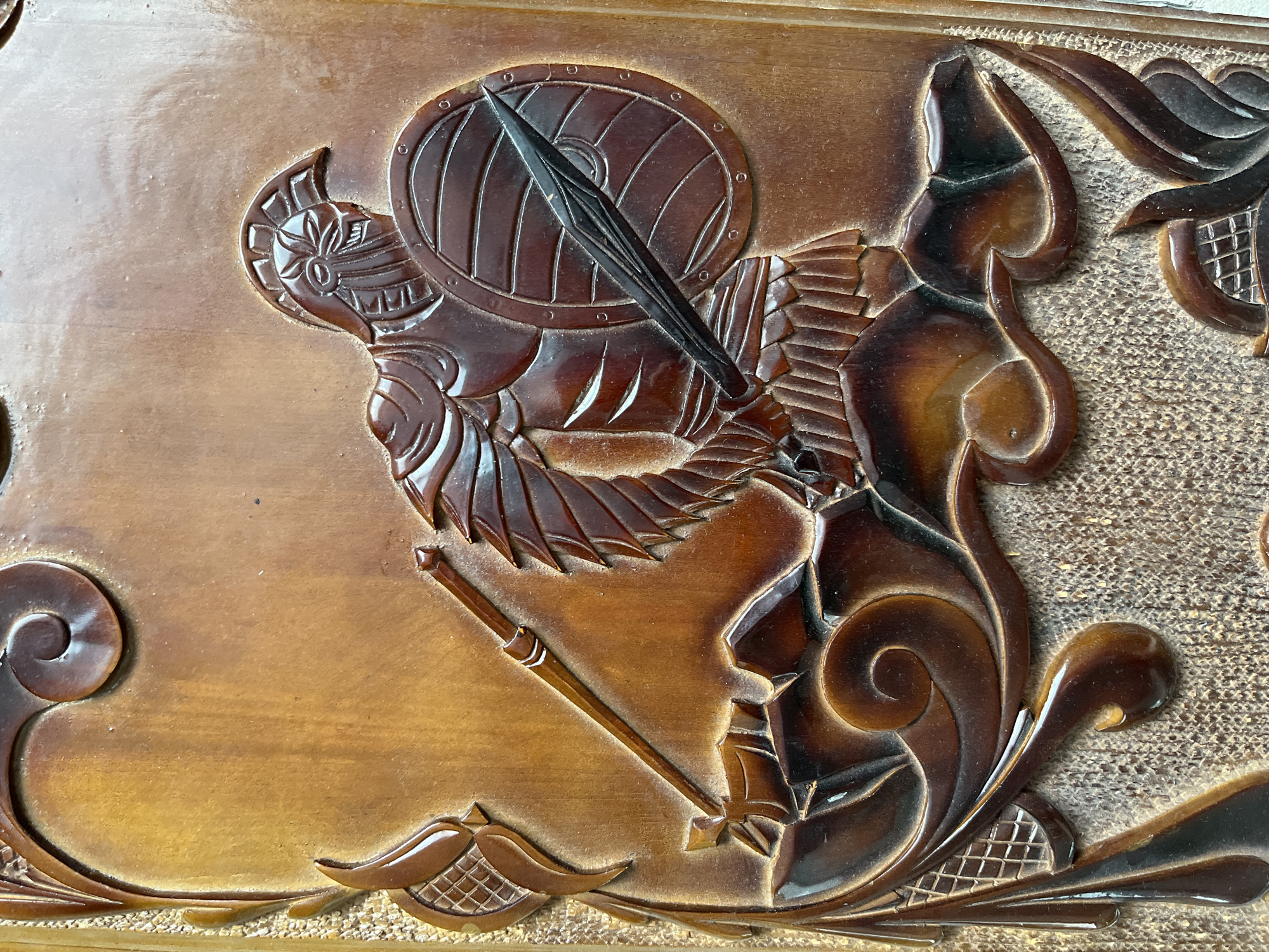 The image is a close-up, top-down view of a detailed wood carving. The carving depicts a figure in a helmet and armor, holding a round shield and a spear. The figure is positioned on a rocky outcrop, with decorative scrolls and floral elements framing the scene. The wood is a warm brown color with a polished finish, highlighting the depth and texture of the carving. The carving is likely part of a larger decorative piece. There is no information in the image that would suggest its location in Saratov, Russia.