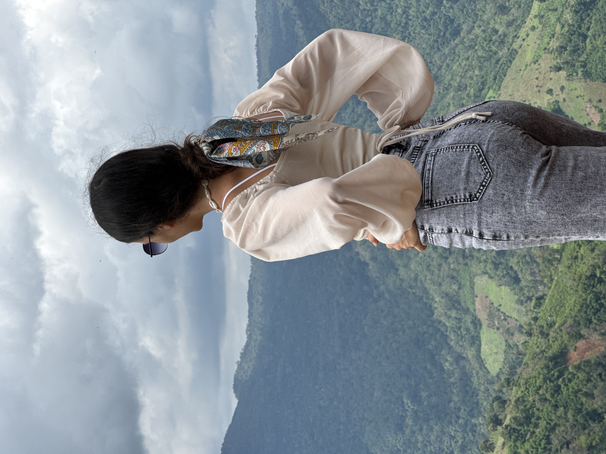 A woman stands on a mountain overlooking a valley in Nao Se, Vietnam. She is wearing a light-colored blouse with puffed sleeves and jeans. Her hair is pulled back in a ponytail with a patterned scarf. Sunglasses are perched on her head. The background is a lush, green, mountainous landscape under a cloudy sky.