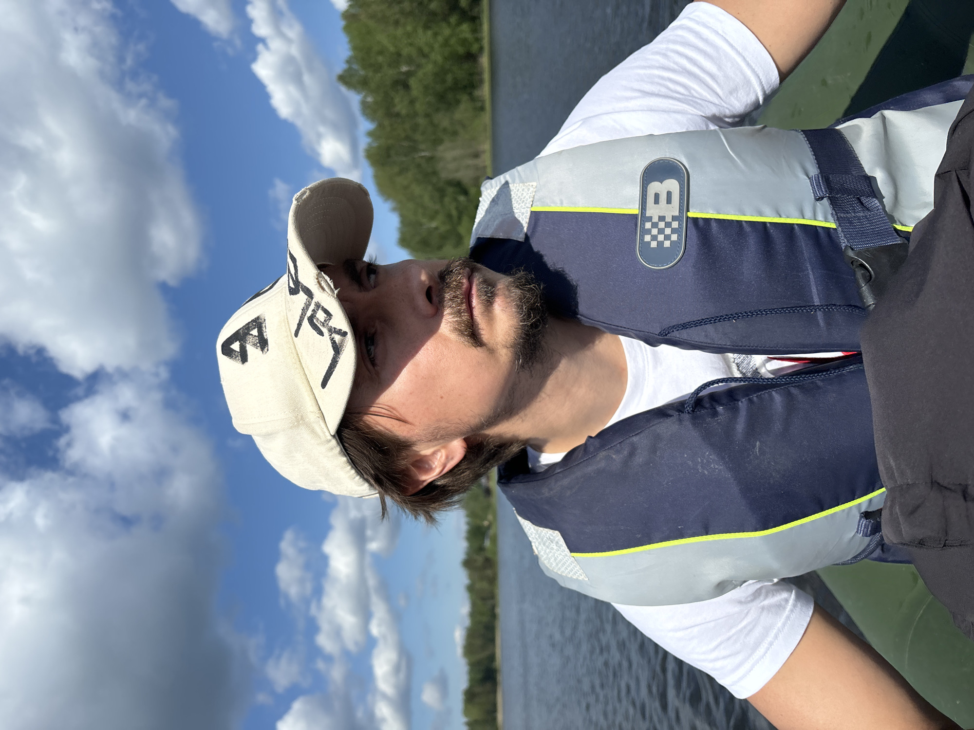A medium shot depicts a person wearing a baseball cap, a white T-shirt, and a life jacket. The life jacket is dark blue and gray with a yellow stripe and a logo containing the letter "B" above a checkered pattern. The person has facial hair and is looking upwards. The background features a body of water, a shoreline with trees, and a sky with clouds. The exact location is Unknown, Unknown.