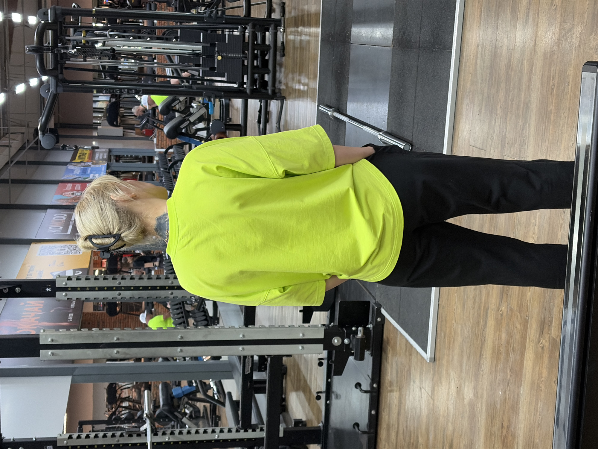 A person with blonde hair and a black hair clip is standing in a gym in Chaikovsky, Russia. The person is wearing a bright yellow shirt and black pants. The gym has weight machines and other exercise equipment. Other people can be seen working out in the background. The floor is a mix of wood and grey tile. A metal bar is near the person's feet. Wall posters are also visible in the background.