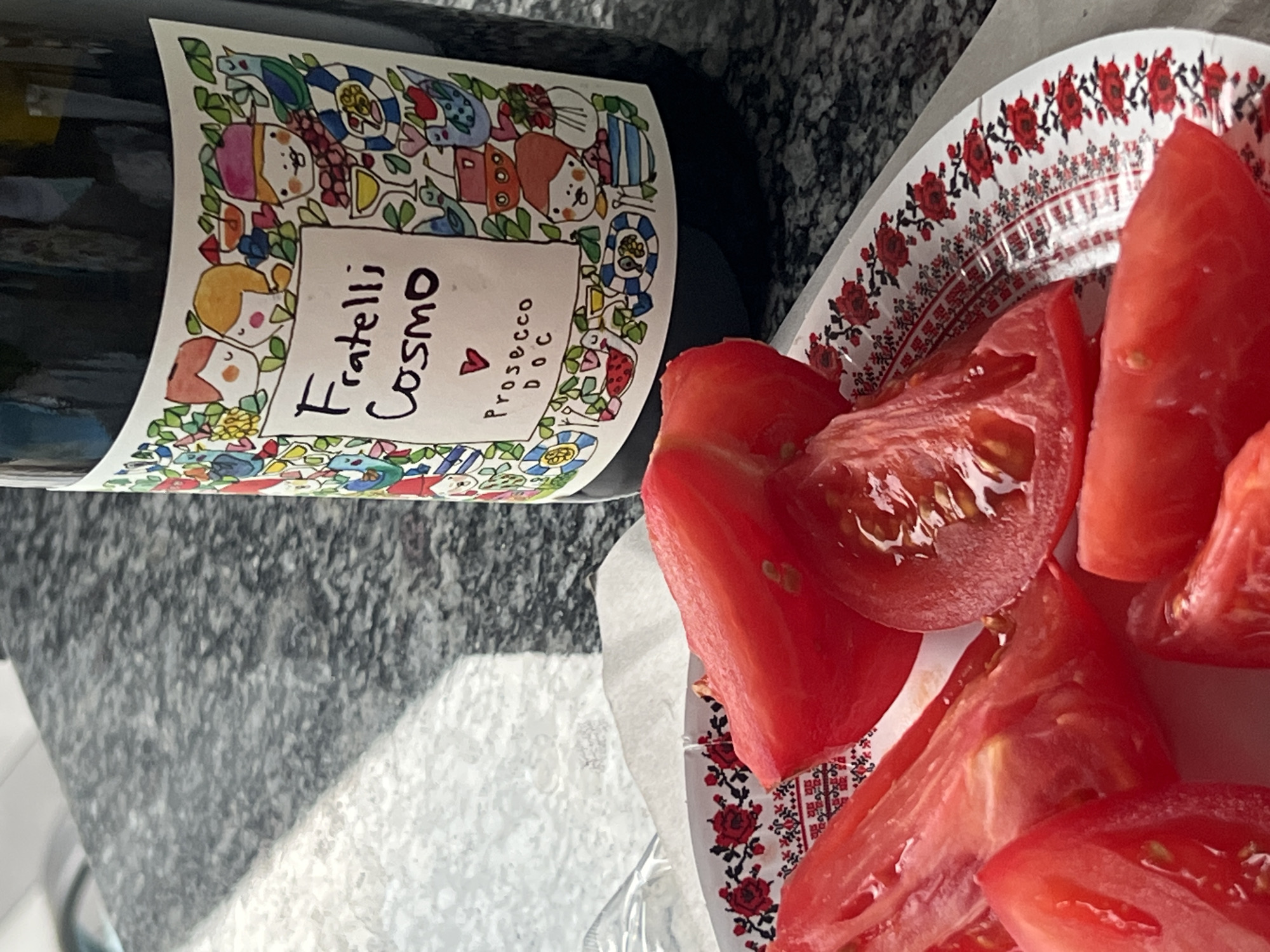 A close-up shot captures a bottle of Fratelli Cosmo Prosecco DOC positioned on a speckled gray countertop. To the left, a white paper plate adorned with a red floral pattern holds several wedges of bright red tomatoes. The bottle's label features a whimsical, colorful illustration of cartoon figures and whimsical designs. The lighting suggests an indoor setting, possibly a kitchen or dining area, with sunlight casting a shadow across the countertop. The overall scene depicts a casual, perhaps culinary, moment.
