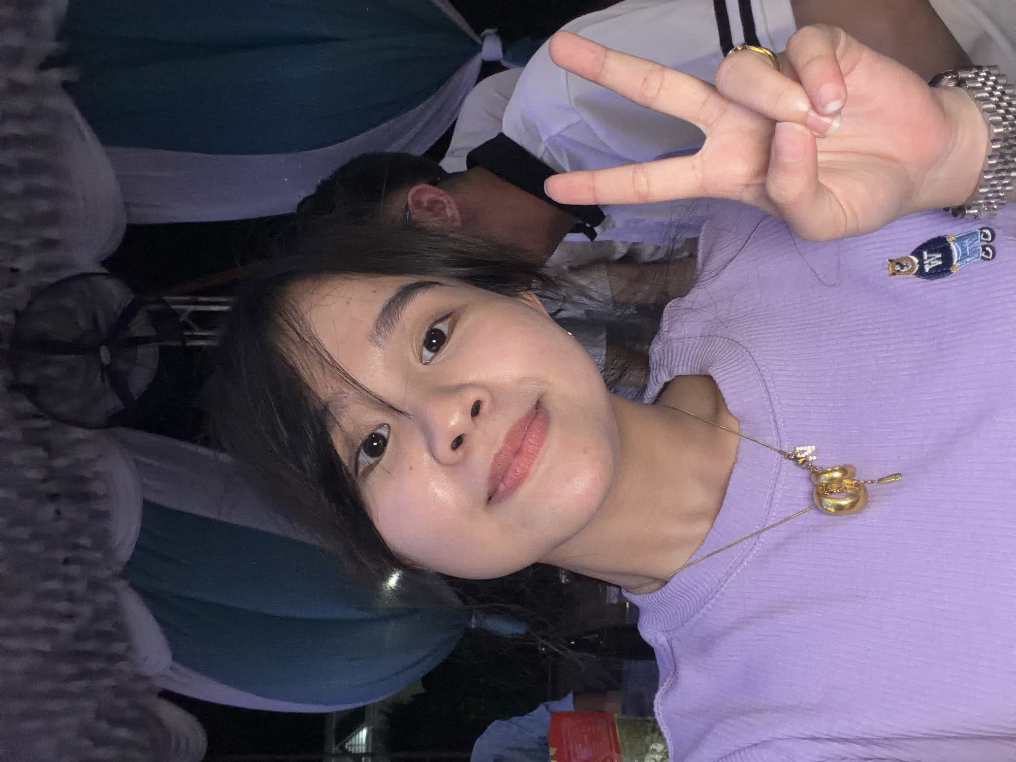 A young woman, likely in her late teens or early twenties, poses for a casual selfie, looking directly at the camera with a slight smile. She has dark hair, styled with bangs, and is wearing a light purple ribbed top. Her left hand is raised, forming a peace sign with her index and middle fingers. A gold-toned ring is visible on her ring finger, and a metallic watch adorns her wrist. Around her neck, she wears a delicate gold-colored chain with an ornate pendant that appears to be intertwined golden rings or a knot. On her shirt, near her right shoulder, is a small embroidered patch or pin depicting a figure in a blue outfit with the letter "W" emblazoned on it.
Behind her, partially visible, is another individual, seen mostly from the back of their head and shoulder, wearing a white shirt with black details. The background is dimly lit, suggesting evening or nighttime, and features draped fabrics in shades of white, light purple, and dark teal, possibly part of a decorative setup for an event or a covered outdoor dining area. A red object, possibly a cup or bucket, is faintly visible in the lower right. The overall setting implies a casual social gathering or outing in a relaxed environment, potentially in Bình An, Vietnam.