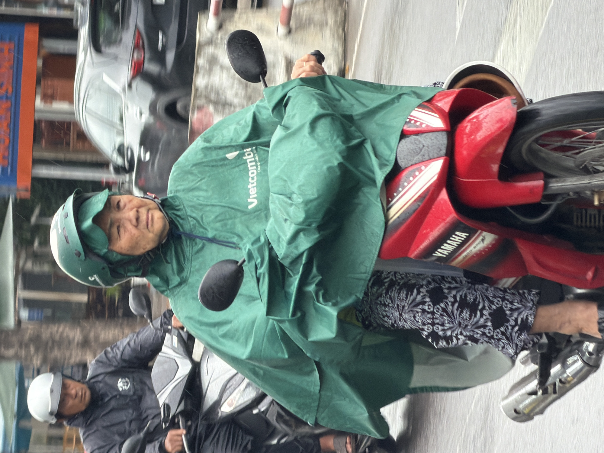 The image captures a rainy day in Nha Trang, Vietnam, with two individuals on motorcycles. In the foreground, a woman wearing a green raincoat and helmet rides a red Yamaha motorcycle. She is wearing patterned trousers and a large green poncho that covers her and the motorcycle. To her left, another person, possibly a man, rides a silver motorcycle, also wearing a dark jacket and a white helmet. They are both moving on a wet road, indicating recent rainfall. In the background, a dark-colored SUV is visible, along with a building with an orange and blue sign that reads "HOÀN SINH" in Vietnamese. The scene is set on a street, likely a busy one, given the presence of multiple vehicles and signage.