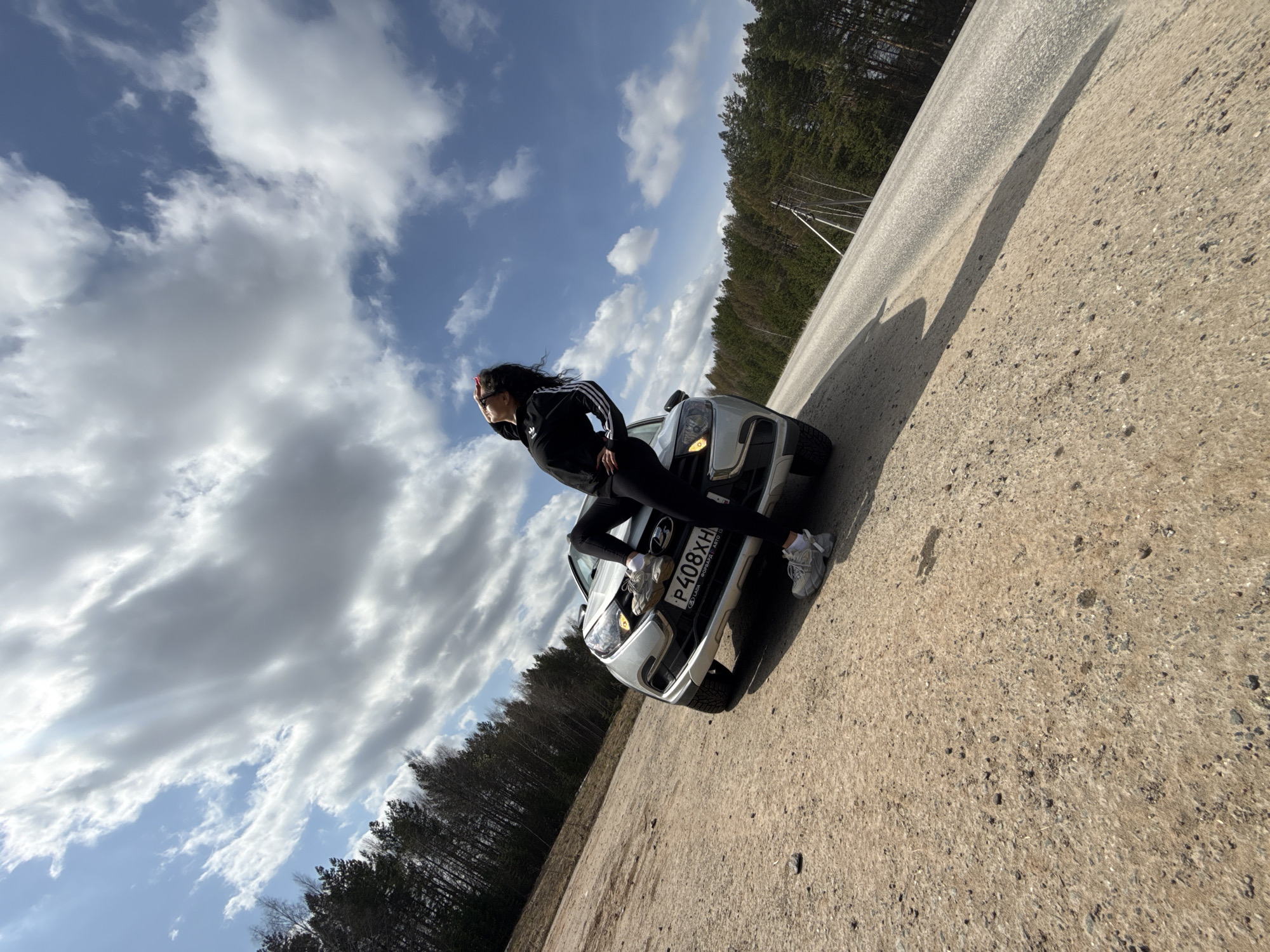 The image features a woman posing on the front of a white car, set on the unpaved shoulder of a rural road in Maksimovo, Russia. The shot is dynamically angled, with the horizon tilted.
The subject is a woman with long, dark, wavy hair, dressed in a black athletic tracksuit comprising a jacket with white stripes on the sleeves and black leggings. She wears light-colored, chunky sneakers. Her right foot is placed on the car's front bumper, and she leans back with her left arm resting on the hood, looking towards her left. Her pose appears confident and relaxed.
The car is a white Lada, confirmed by the visible text "LADA DORAB.RU" below the front license plate. The license plate clearly reads "P408XH 190".
The scene unfolds during bright daytime, with direct sunlight casting distinct shadows on the ground. The sky above is a mix of clear blue patches and large, white and grey clouds. A dense forest of coniferous trees lines the road in the background, with some utility poles visible among them. The road transitions from an unpaved shoulder to an asphalt surface further away.