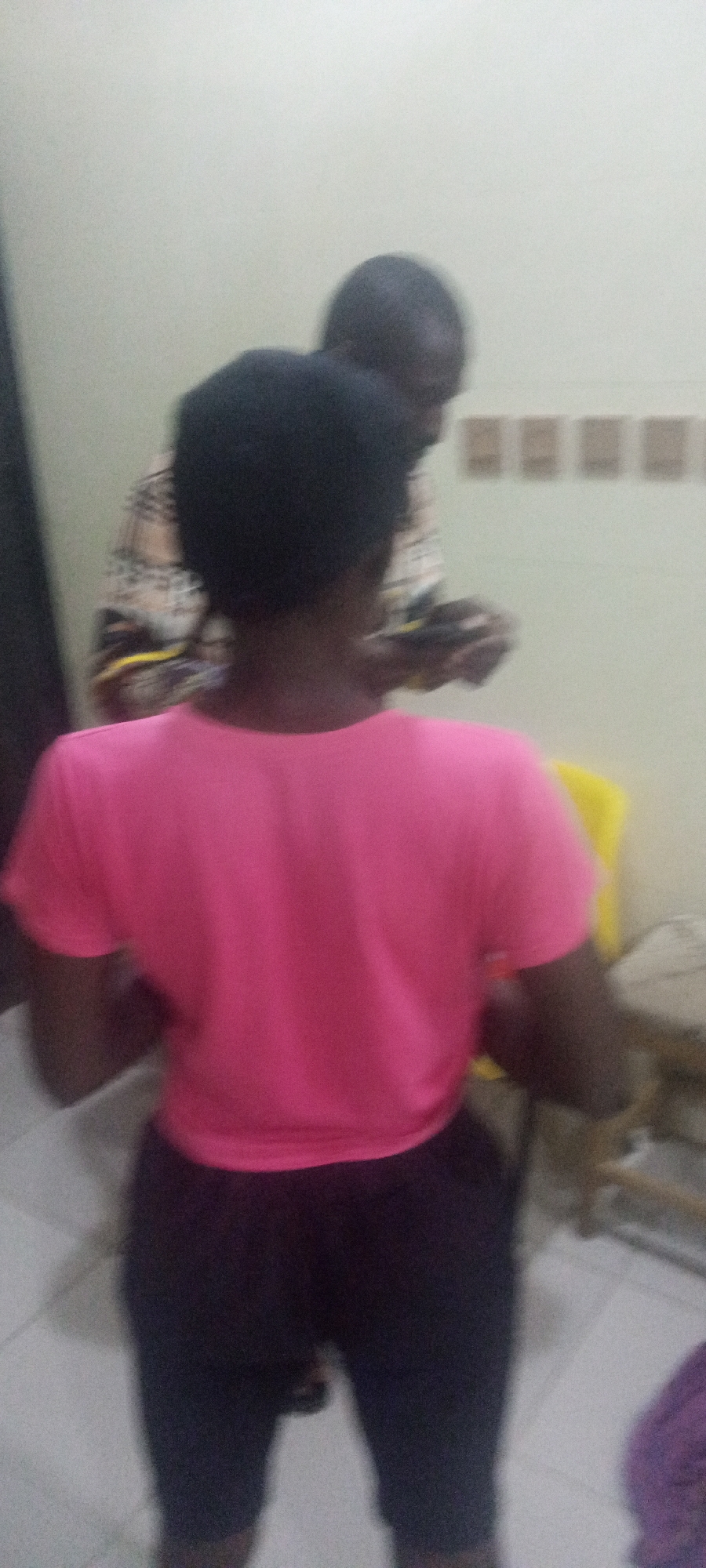 Two individuals are present. One individual is in the foreground, facing away from the camera, wearing a pink shirt and dark pants. Behind this individual stands another person, slightly blurred and in the background. This second person appears to be using a mobile device. The setting appears to be indoors, with a wall visible and a few furniture items in the periphery. The specific location is Unknown, Unknown.