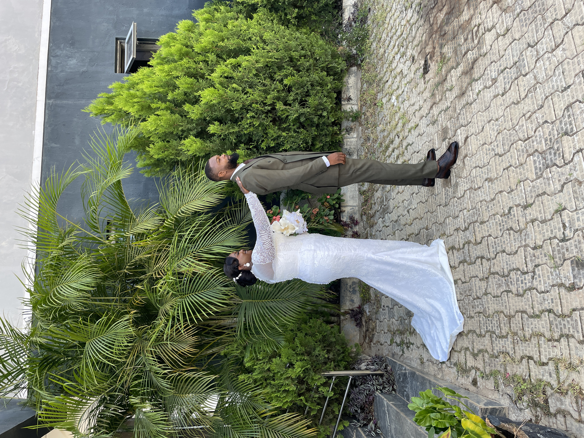 A bride and groom pose in front of a building with shrubbery, likely in Jos, Nigeria. The groom, dressed in a brown suit and brown shoes, stands on a paved path. The bride, in a long white gown, raises her arm towards his face, holding a bouquet of white flowers. Lush green plants and palm trees surround them, and a grey building with a window is visible in the background.