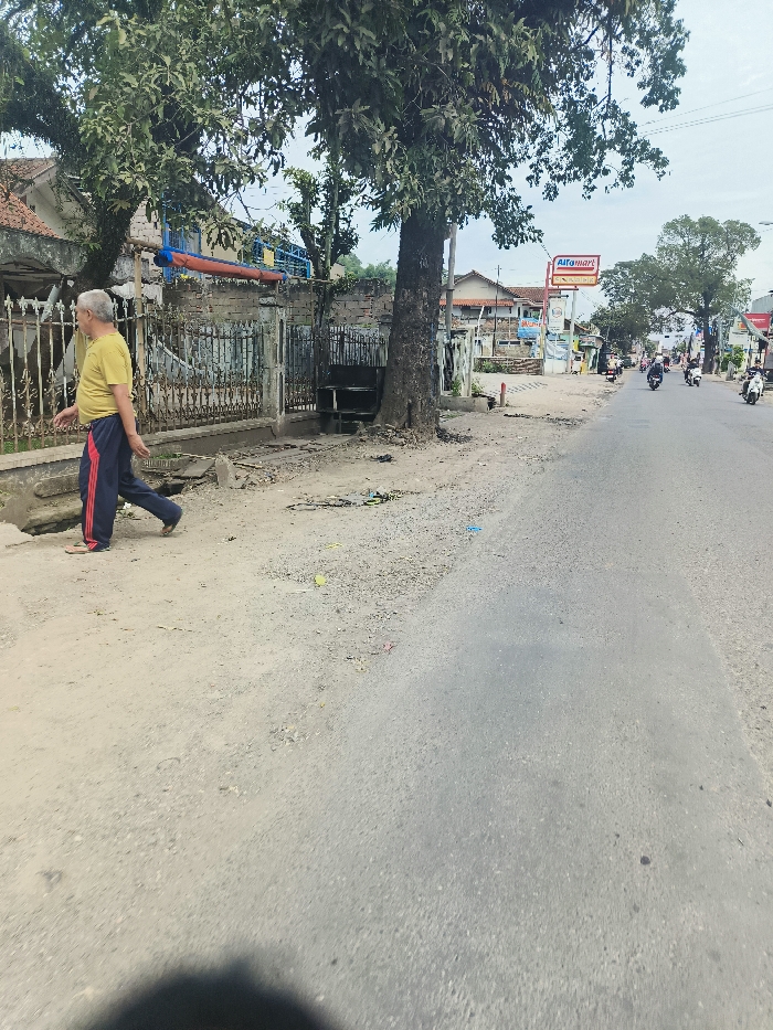 The image shows a man in a yellow shirt and red-striped pants walking on a street in Ciparay, Indonesia, at coordinates -7.025 latitude and 107.69 longitude. The street is paved and there are several buildings on both sides of the street. There is a large tree on the left side of the image, and several motorbikes are parked on the right side of the image. The sky is cloudy.