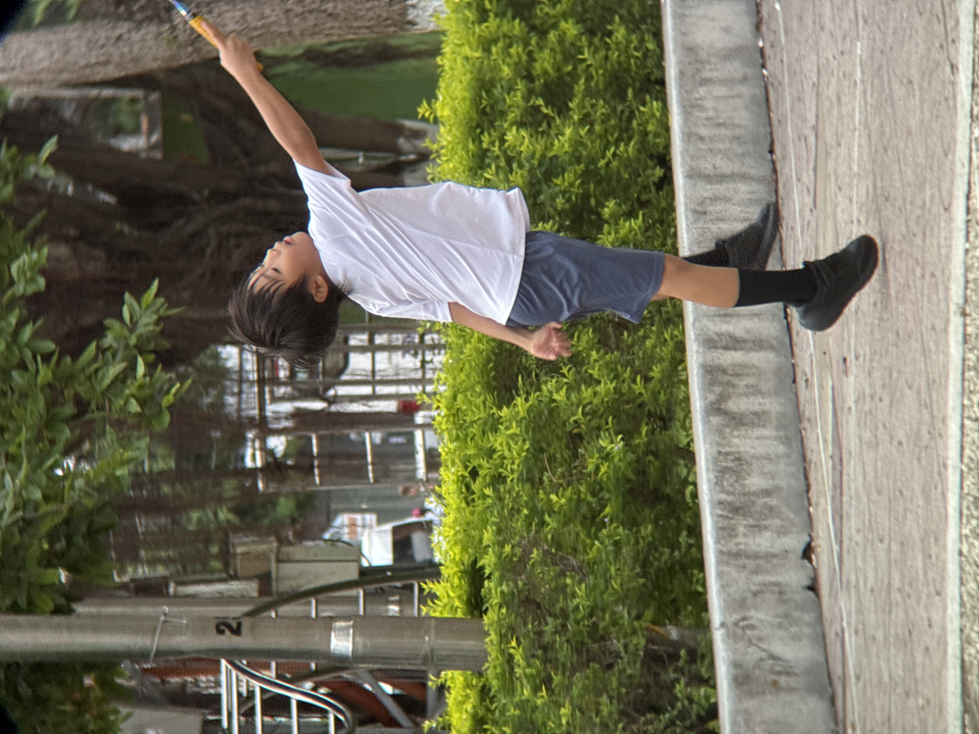 A young boy is playing badminton in what appears to be a public park or recreational area. He is wearing a white t-shirt, dark blue shorts, black socks, and black athletic shoes. He is captured mid-air, appearing to be in the process of striking a shuttlecock with his racket, which is partially visible. The background consists of lush green bushes, a concrete wall, a paved ground surface, and trees. In the far background, there are hints of urban structures, including a lamppost with the number "2" on it, a railing, and a white vehicle. The lighting suggests it is daytime, with soft shadows. The overall setting is consistent with a public space in Nha Trang, Vietnam, likely an outdoor sports facility or park.