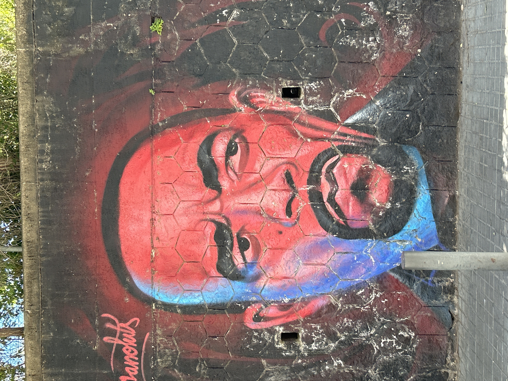 A mural depicting a face painted in red, black, and blue adorns a concrete wall composed of hexagonal blocks. The face is stylized with heavy black outlines around the eyes, nose, and mouth. The background is predominantly black with hints of red. The wall appears weathered with some deterioration and white staining. At the bottom left, there is a signature or inscription in cursive script. The wall is located in the city of l'Hospitalet de Llobregat, Spain. Adjacent to the right side of the mural is a tiled sidewalk with a metal pole extending upwards. Some greenery is visible on the left edge of the mural.