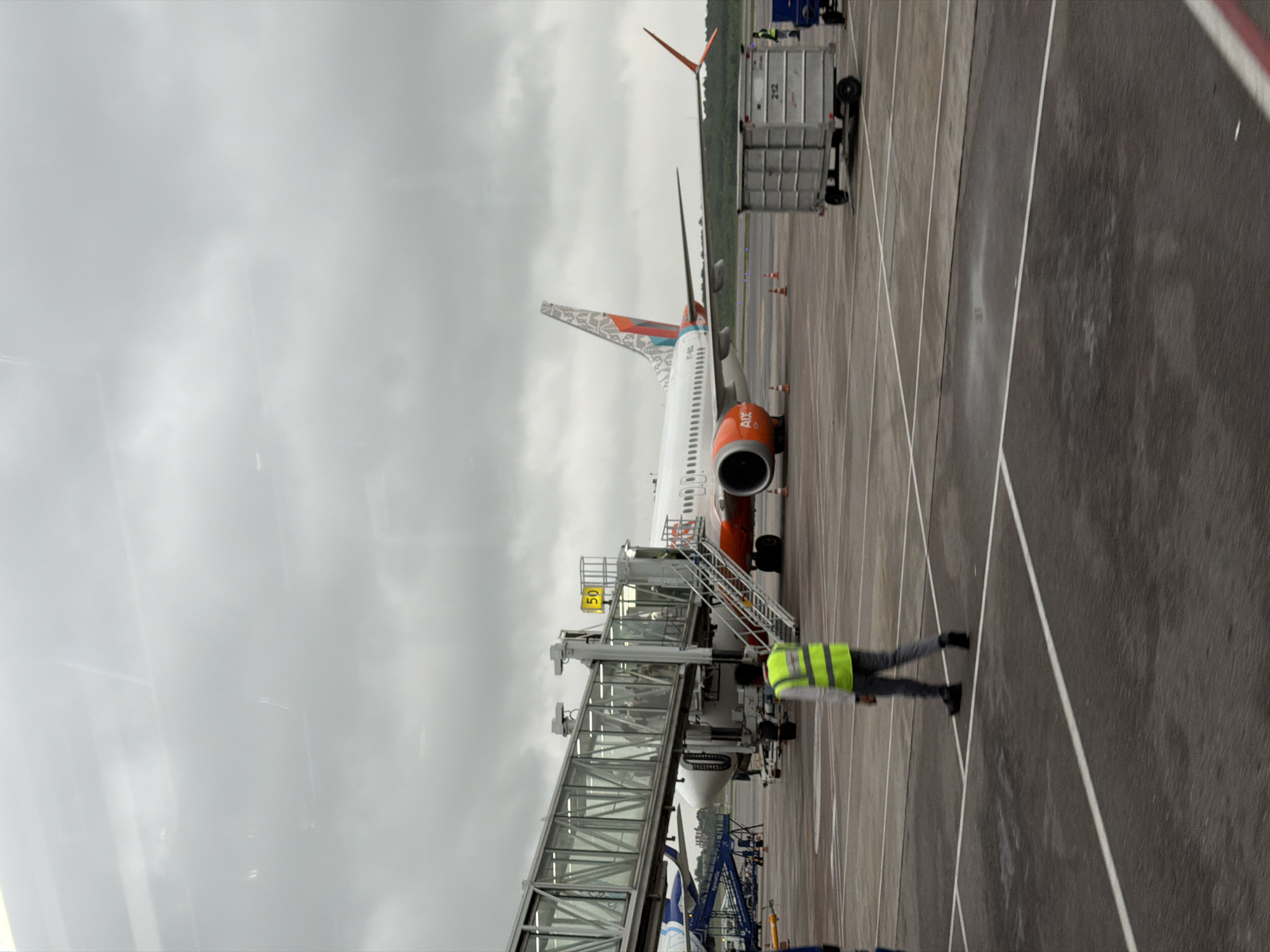 The image shows an airport tarmac under an overcast sky. A jet bridge is extended to an airplane, which has "AIX" visible on its engine. A person wearing a high-visibility vest walks away from the viewer on the tarmac. In the background, a luggage cart is parked near the airplane, and another small blue vehicle is to its right. The scene appears to be at Rajiv Gandhi International Airport in Hyderabad, India, indicated by the presence of an AIX Connect (formerly Air India Express) aircraft and common airport infrastructure. The tarmac is marked with white lines, and there are some orange cones near the aircraft.
