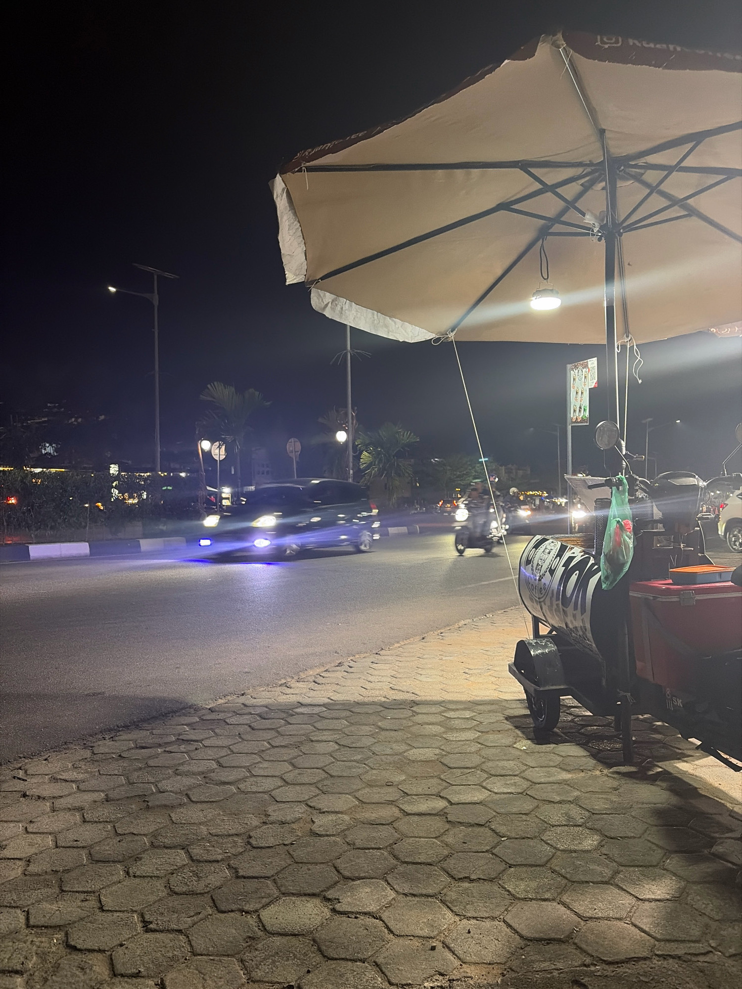 The image captures a nighttime scene on a city street in Tanjung Pinang, Indonesia, likely in a commercial or entertainment district. A large, beige umbrella is prominently featured in the foreground, casting a shadow and illuminating a small hanging lamp. Beneath the umbrella, a metal cart with wheels is parked, adorned with black and white text. To the left of the cart, a red cooler is visible. A green plastic bag hangs from the cart. The street itself is paved, with hexagonal stone tiles forming the sidewalk in the foreground. Across the street, vehicles, including cars and motorcycles, are in motion, their headlights and taillights creating streaks of light. The background shows a row of trees, streetlights, and buildings with illuminated windows, suggesting an active urban environment. The overall impression is that of a street-side establishment or vendor operating late into the night.