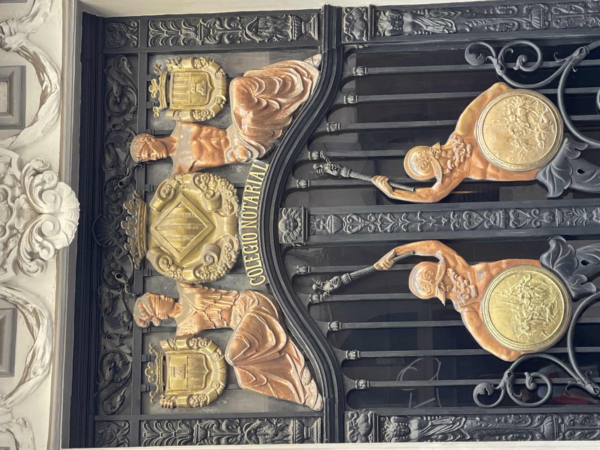 This detailed image captures the ornate metalwork entrance of the Colegio Notarial de València, located in València, Spain. The scene depicts a multi-layered, decorative gate structure.
The upper section features a bronze and gold tableau. Centrally positioned is a golden rhomboid emblem, flanked by grapevine motifs, with an indistinguishable symbol within, and underscored by the curved golden text "COLEGIO NOTARIAL." To the left of this central emblem is a seated female figure, rendered in bronze or copper tones, with a blindfold covering her eyes (depicting Lady Justice), holding a golden shield bearing a single tower with battlements. To the right is a seated, bare-chested male figure, also in bronze or copper, holding a golden shield displaying a tower with battlements and vertical stripes. Both figures are situated against a background of dark, intricately carved metalwork featuring floral and scroll patterns, beneath a light-colored, ornate architectural molding.
The lower section comprises the functional gate, crafted from dark, possibly wrought iron. Each of the two gate panels features a large, standing bronze or copper figure. The left figure, a bearded male warrior wearing a helmet, is depicted raising a weapon resembling a mace or club in his left hand, while holding a prominent circular golden shield in front of him. The shield bears a detailed bas-relief scene of multiple figures engaged in what appears to be a historical event or battle. This figure faces towards the right. Symmetrically, the right figure, also a bearded male warrior in a helmet, raises a similar weapon in his right hand and holds an identical circular golden shield with the same relief scene, facing towards the left. The gate bars are vertical, interspersed with decorative elements including small cherub faces, ram heads, and stylized female figures on the vertical support columns, all in dark metalwork. The background seen through the gate is dark, with indistinct reflections.