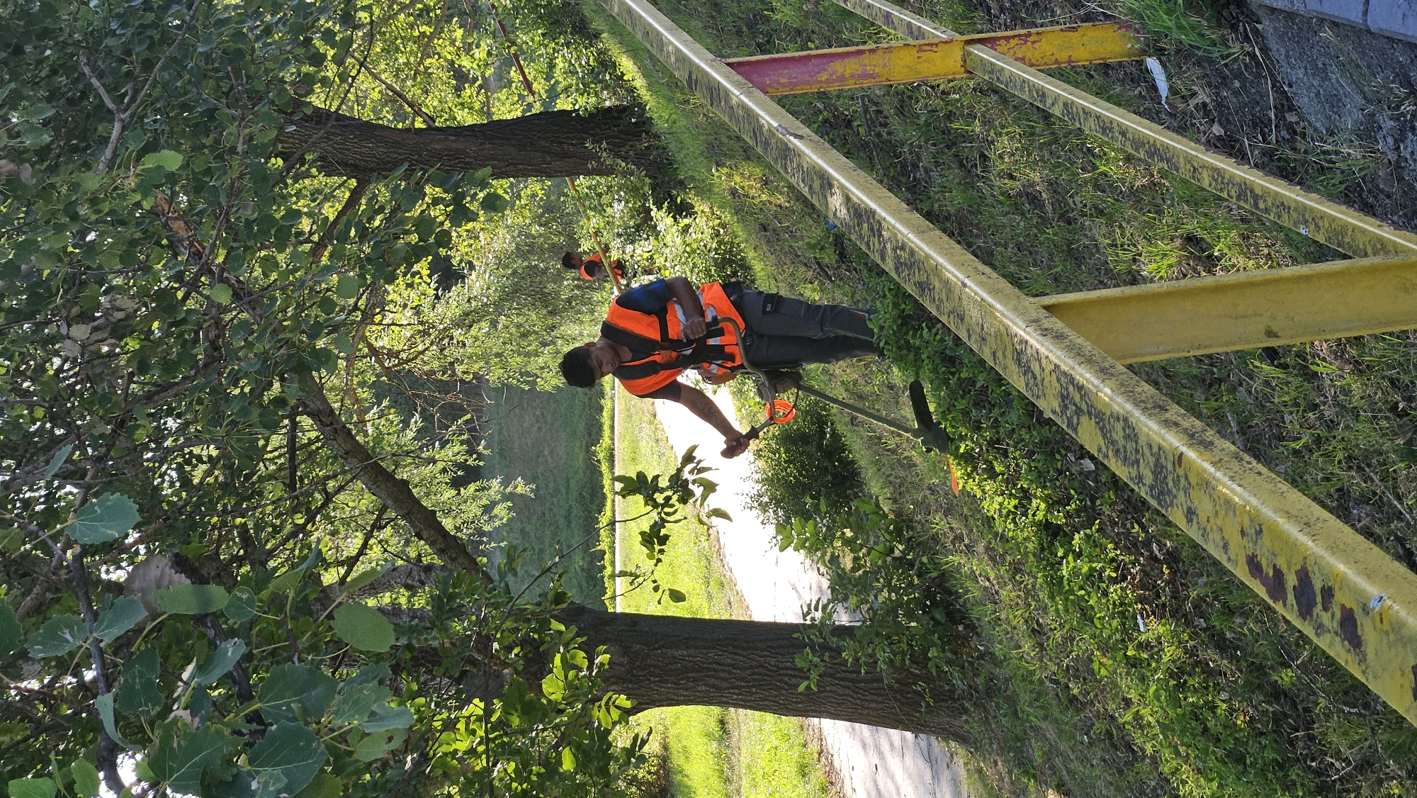 The image shows a man operating a string trimmer on a grassy slope next to a pathway. He is wearing an orange safety vest and dark pants. The slope is covered in green grass and vegetation, with trees in the background. A yellow railing runs along the foreground. In the background, another person in an orange safety vest can be seen further down the slope. The scene appears to be outdoors during daylight hours.