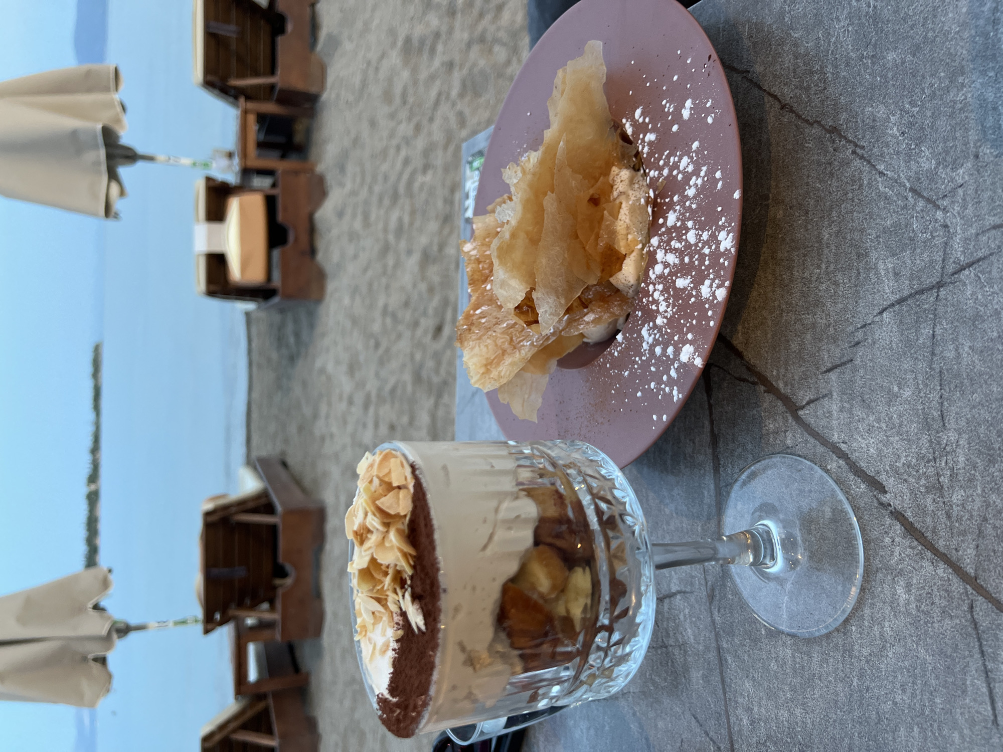 A close-up shot features two desserts on a stone table at the beach in Eretria Municipal Unit, Greece. In the foreground, a glass goblet holds a tiramisu dessert topped with cocoa powder and sliced almonds. To the right, a pink plate displays a dessert with crispy pastry pieces scattered over a creamy base, dusted with powdered sugar. In the background, the blue sea stretches towards a distant shoreline under a hazy sky, with two lounge chairs and a closed umbrella suggesting a tranquil beach setting. The overall scene captures a leisurely moment by the sea.