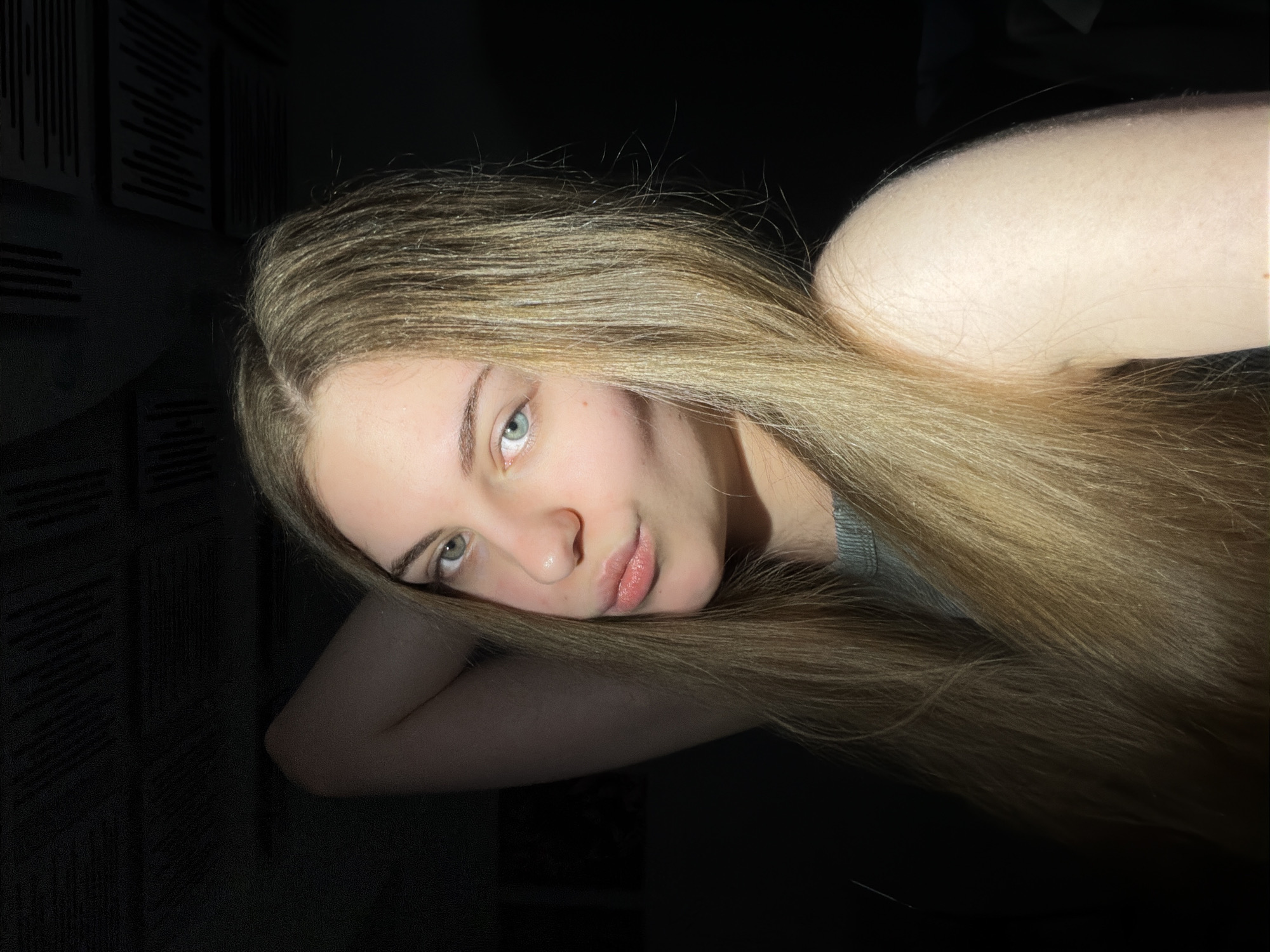 The image shows a young Caucasian woman with long, blonde hair and light blue eyes. She is posing with her arm behind her head and her lips slightly pouted. The lighting is dramatic, with a strong light source creating shadows. The background is dark and appears to be an indoor setting. There is no indication of the location being in Dnipro, Ukraine.