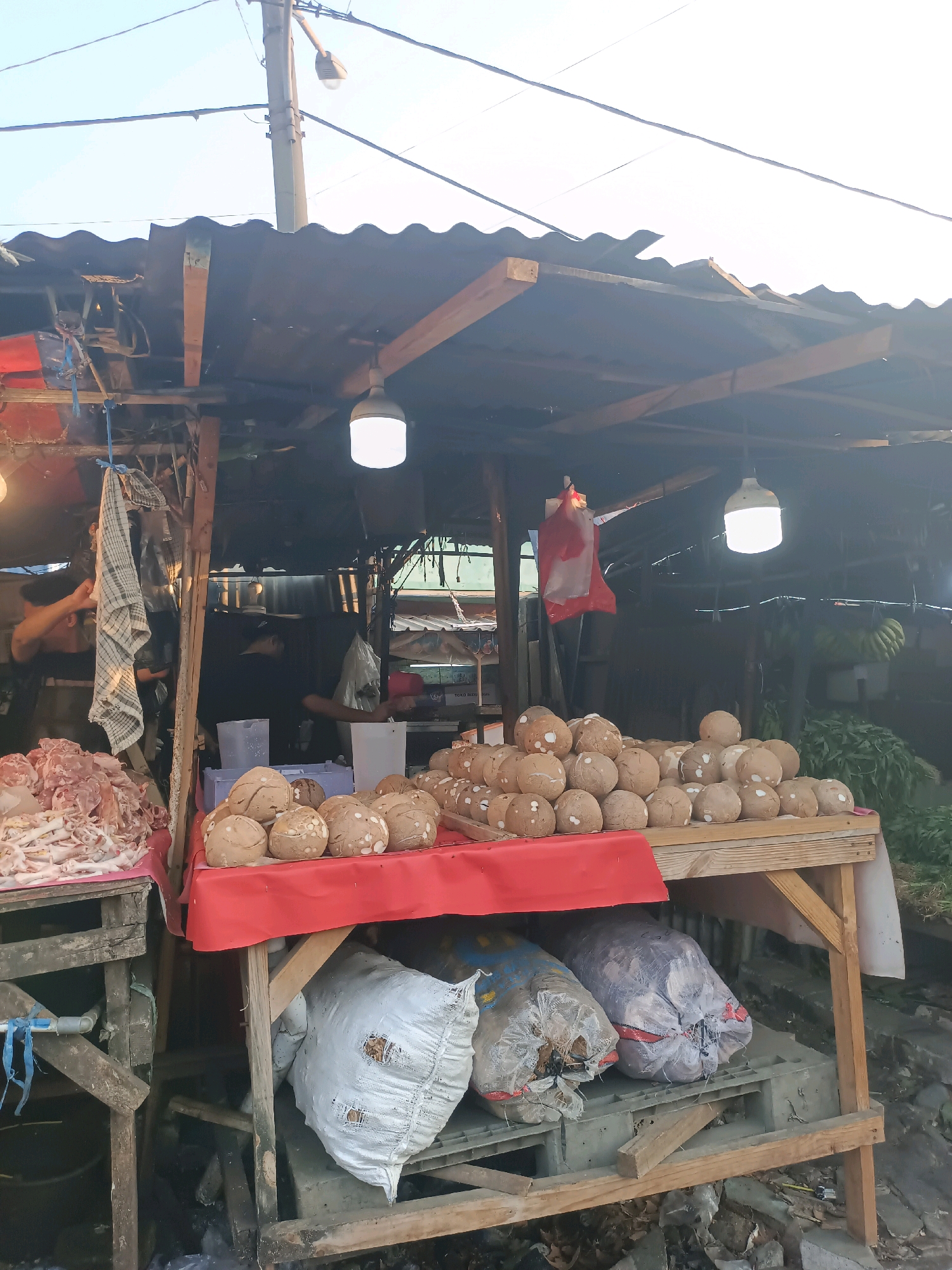 This is a forensic, matter-of-fact description of an outdoor market stall in Karawang, Indonesia. The stall is covered by a corrugated metal roof supported by wooden beams. Two bright LED lights hang from the roof, illuminating the scene.
In the foreground, a wooden table is draped with a red cloth. On this table, a large pile of coconuts is displayed. To the left of the coconuts, on a separate, lower wooden surface, is a pile of raw meat, including what appears to be chicken feet.
Beneath the table, three large sacks are visible. The sack on the left is white and appears to be made of woven material, with two visible holes. To its right, there are two more sacks, one blue and one grey, also appearing to be made of woven material and filled with what could be produce or other market goods.
In the background, two individuals are visible behind the counter. One person on the left, wearing a black shirt, is partially obscured as they interact with the meat display. Another person, also in dark clothing, is visible further back. Various market stalls and produce, including what appear to be bunches of bananas and leafy greens, are discernible in the background, suggesting a bustling market environment. The ground around the stall is covered with debris and appears to be unpaved. Power lines and a utility pole are visible overhead, indicating the presence of public infrastructure.