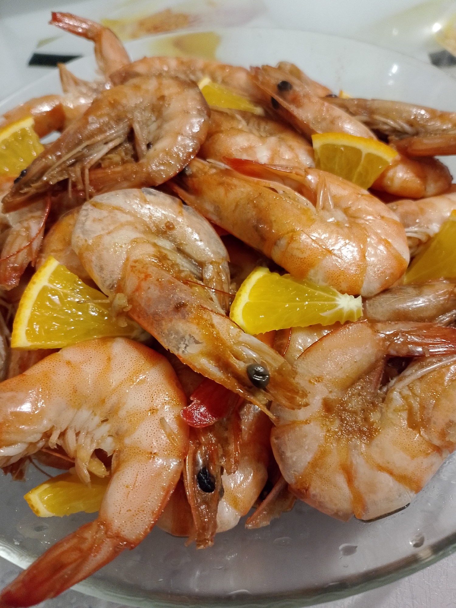 A close-up shot depicts a serving of cooked shrimp, interspersed with wedges of orange. The shrimp exhibit a cooked, reddish-orange hue, and some have visible antennae. The scene is set in a bowl, possibly made of glass, on a light-colored surface. There is no indication about the city of Noviy or Russia in the image.