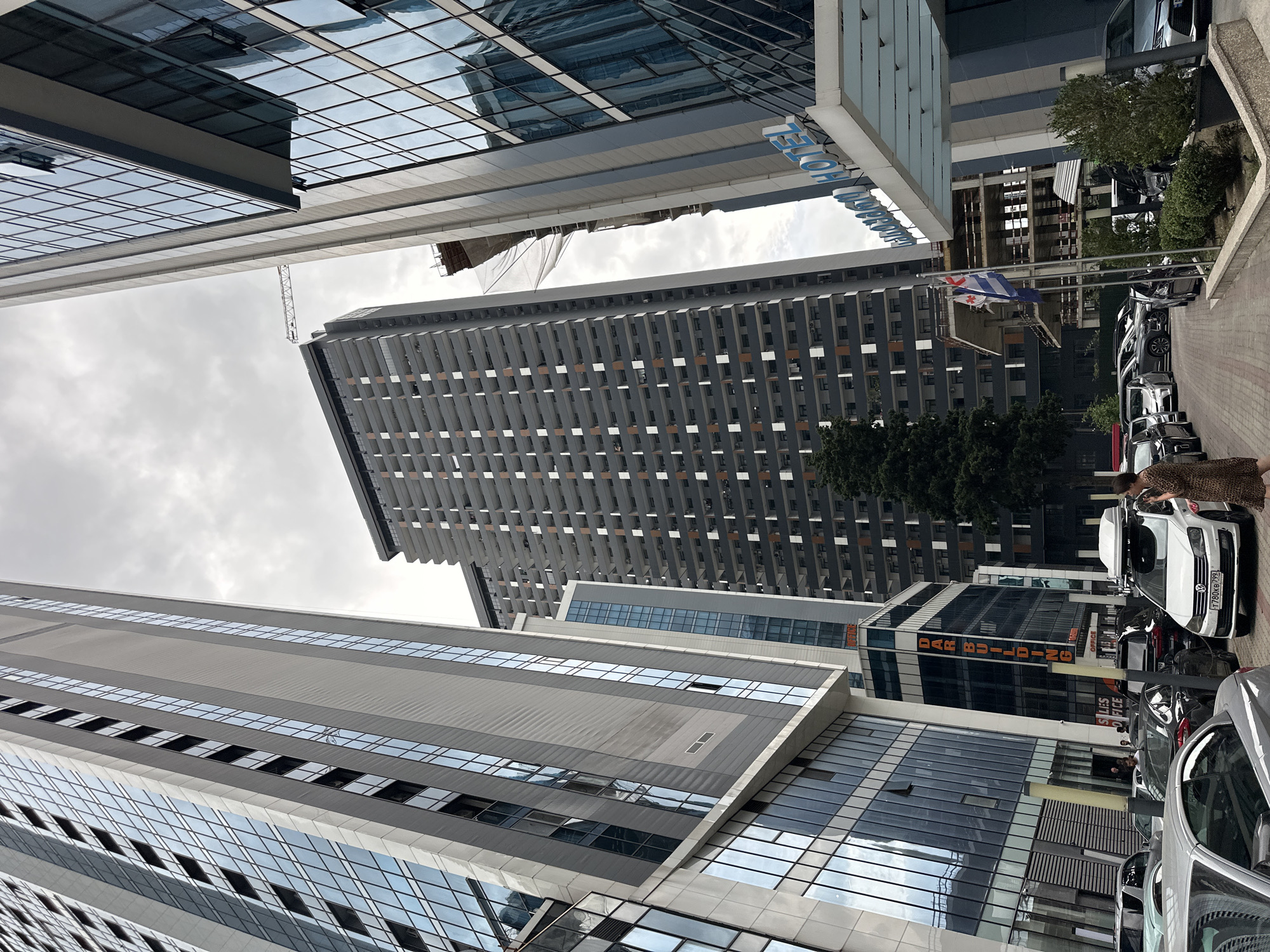 The image shows an urban scene in Batumi, Georgia, featuring modern architecture and a street with parked cars. Several tall buildings dominate the frame, characterized by their glass facades and geometric designs. One building has a blue sign that reads "HOTEL" in large letters. In the background, a crane suggests ongoing construction.
Several cars are parked along the side of the street. One white car, a Volkswagen, has a license plate with the text "T 780KG79". A woman wearing a leopard-print dress is standing next to the white car, seemingly interacting with its interior. Other vehicles, including darker cars and what appears to be a silver sedan in the foreground, are also visible.
Two national flags are displayed on poles between buildings; one is the flag of Georgia and the other appears to be the flag of Ukraine. The sky is overcast, indicating a cloudy day. The overall impression is that of a typical day in a city, with a focus on commercial buildings and street-level activity. Visible text includes "HOTEL", "T 780KG79", "DAR BUILDING", and "SALES OFFICE".