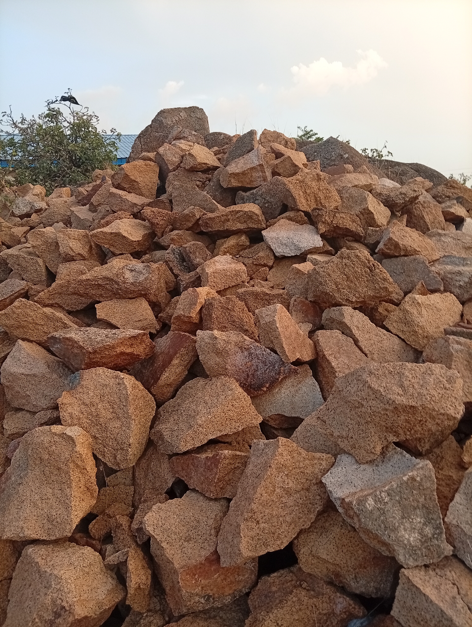 The image shows a large pile of irregularly shaped, brownish rocks. The rocks are stacked haphazardly, with varying sizes and textures. In the background, there's a hint of foliage, possibly trees or bushes. There is also an object, possibly a bird, perched on top of this vegetation. The sky is partially visible, exhibiting a mix of light blue and whitish tones, with a few clouds. The image is in focus, revealing the details of the rocks. The location is in Jos, Nigeria.