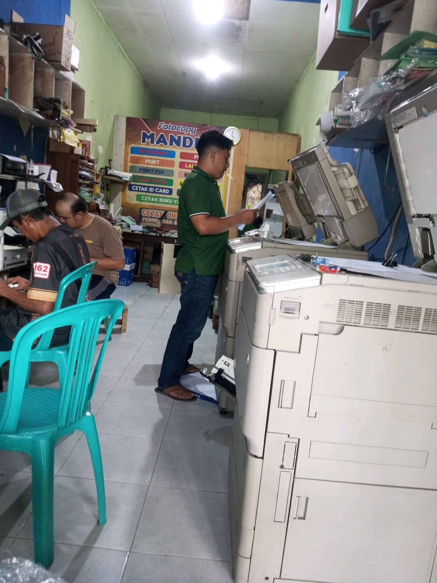 The image depicts the interior of a small photocopy shop, possibly located in Garung, Indonesia. In the foreground, a teal plastic chair is visible. To the left, two men are seated, one wearing a grey baseball cap and a black t-shirt with "162 ECTION" printed on it, and the other a brown t-shirt. In the mid-ground, a man wearing a green polo shirt and blue jeans stands and operates a large, beige photocopier. He is holding a document in his hands. Behind him, a banner with "Fotocopy MANDIRI" is displayed on the wall. The shop appears to be filled with office equipment, including shelves stacked with boxes and paper. The lighting is somewhat dim, with a bright light fixture visible on the ceiling. The floor is tiled.