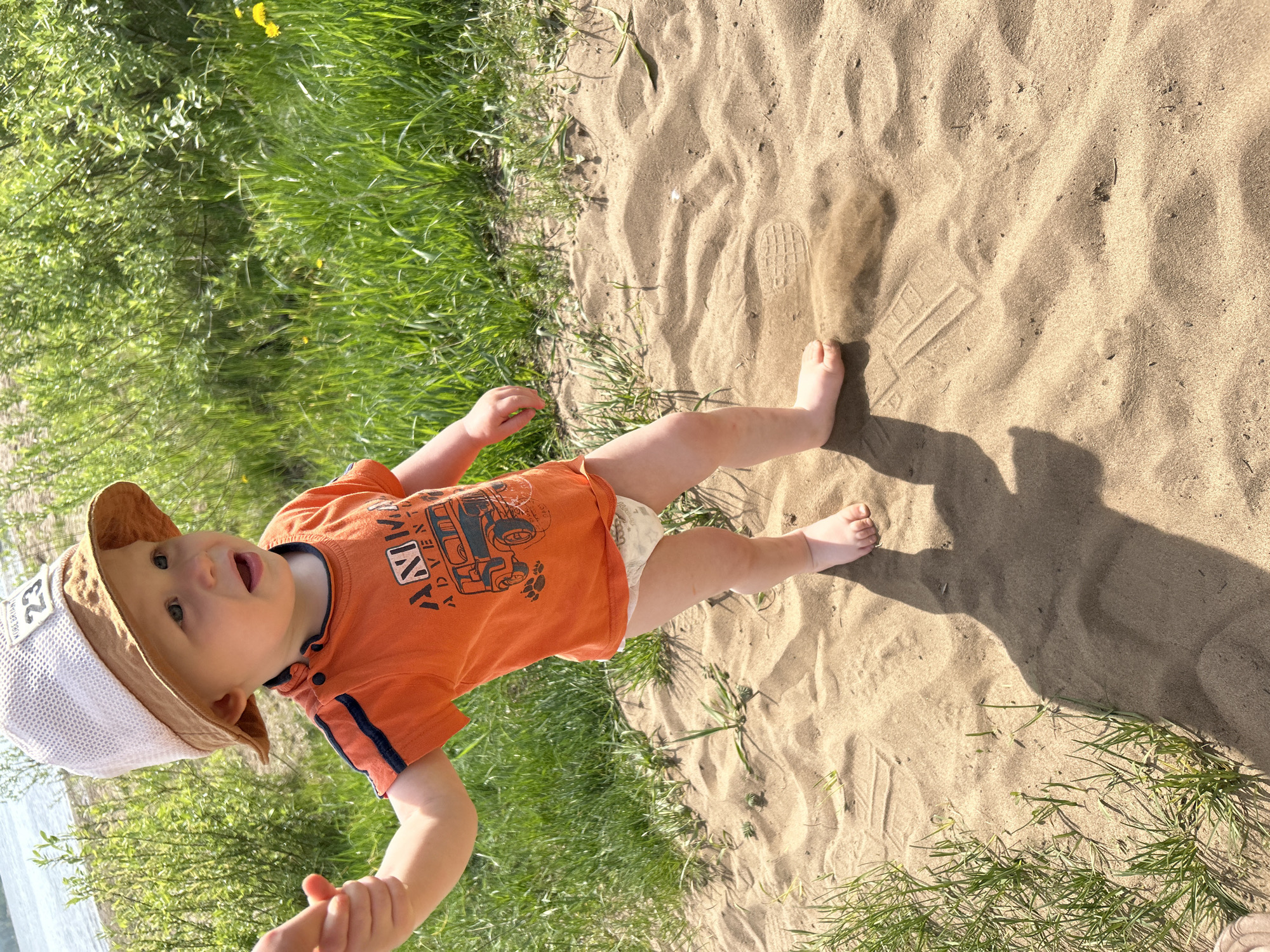 A toddler stands on a sandy beach next to a grassy area. The toddler wears an orange shirt with a truck design, a diaper, and a bucket hat. The background includes green vegetation and a body of water, possibly a river or lake. There are footprints in the sand. The location is likely near a beach area or park in Chaikovsky, Russia, based on the image's origin information.