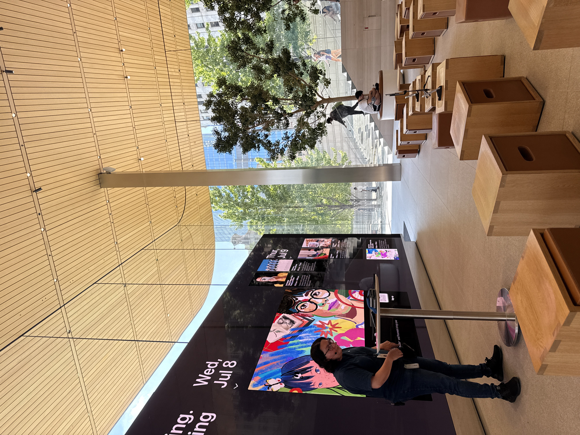 This is a full-shot, eye-level, outdoor photograph taken at the Apple Store Chicago. The store's interior features a large, curved, wooden slatted ceiling and a wall with a large screen displaying colorful artwork and event information. A person wearing a dark shirt and jeans, with glasses and a headset, stands in front of the screen, facing a small table with a tablet.
The store's exterior is visible through a large glass wall, showing a staircase leading to an upper level, trees, and other buildings in the background. People are seen walking up the stairs and sitting on benches. In the foreground, there are several wooden cube-shaped seats. The overall impression is a modern, open-concept retail space with a blend of indoor and outdoor elements.
