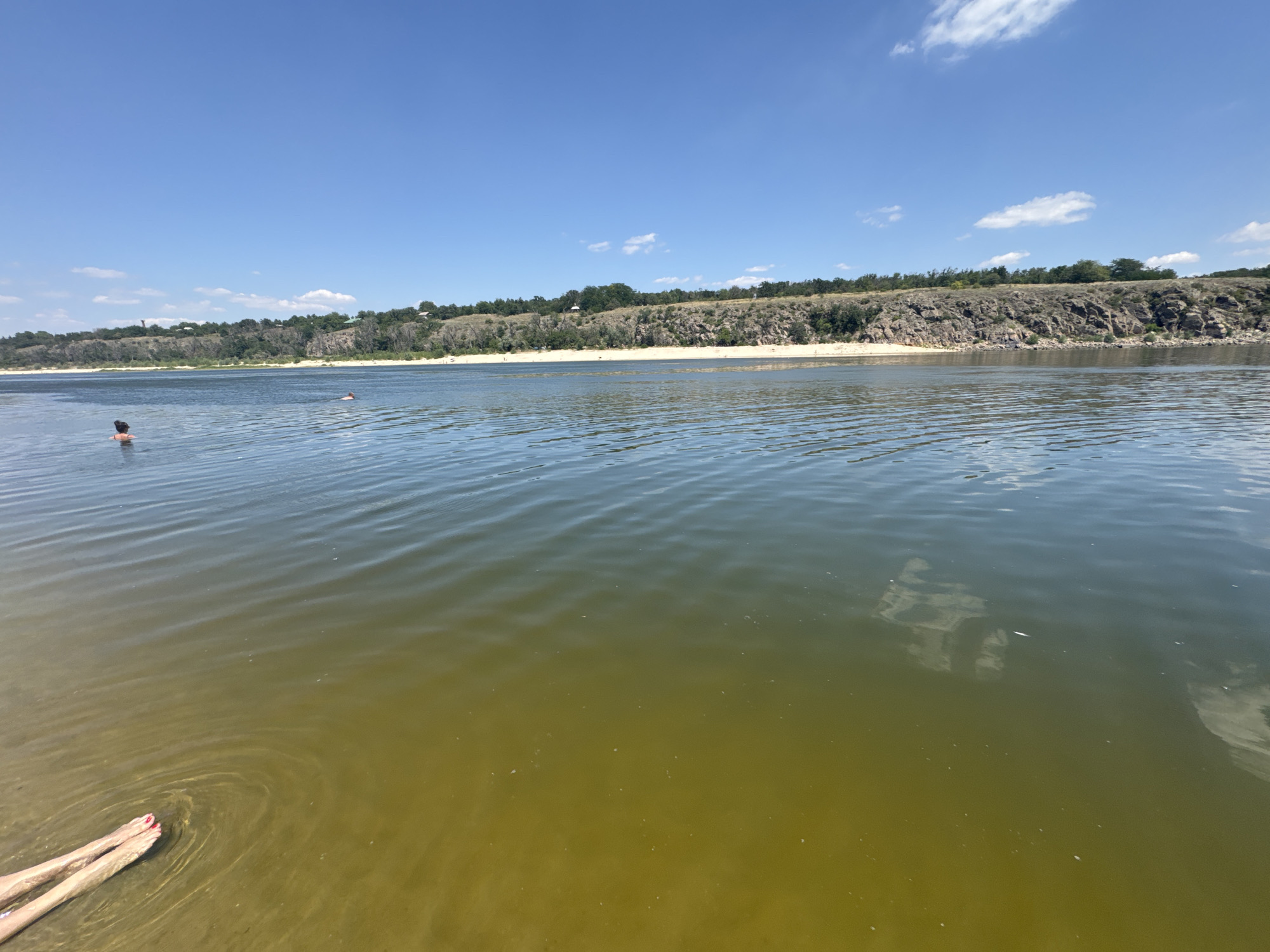 The image captures a wide, tranquil river under a clear blue sky with scattered white clouds. The foreground shows a sandy bank and shallow, greenish water where a person's legs, adorned with red nail polish, are submerged. Ripples emanate from the submerged legs. Further into the water, another individual is visible, seemingly swimming. The middle ground features the river's surface reflecting the sky and distant landscape. The background is dominated by a natural embankment with rocky outcrops and lush green vegetation, suggesting a natural or semi-natural park setting. The scene is likely the Dnipro River in Zaporizhzhia, Ukraine, given the distinctive embankment and the presence of a large river.