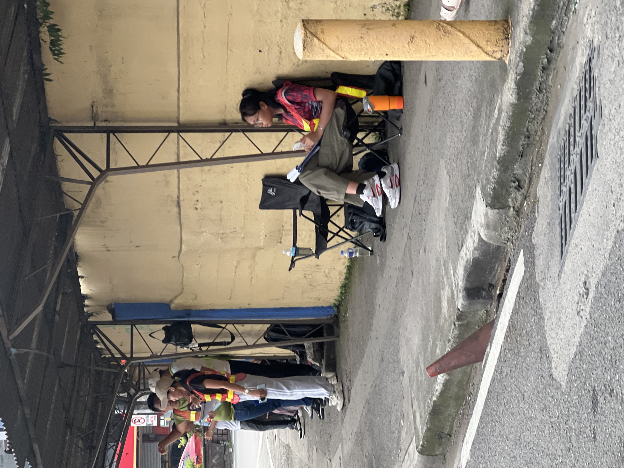 The image shows a street scene in Davao City, Philippines, likely during the daytime. The perspective is from above, looking down at an angle, and the image is rotated.
The primary subject is a young woman sitting on a folding chair under a covered walkway. She is wearing a red and pink tie-dye shirt and olive green pants, with white sneakers. She appears to be looking at her phone or a small device. Next to her are several items, including a black bag, an orange object that resembles a water gun or toy, and two plastic water bottles. There are other folding chairs and bags in the vicinity, suggesting a group or an event is taking place.
In the background, further down the street, a group of people are standing. Some of them are wearing brightly colored vests, possibly indicating they are event staff or participants. One person in the group is looking towards the camera. There are structures that suggest a covered market or waiting area. A red sign with a circular symbol and a line through it is visible, which is a common traffic or prohibition sign.
The ground is paved with asphalt, and there is a drainage system visible along the edge of the walkway. The overall atmosphere appears to be casual, possibly a break during an event or a gathering. The lighting suggests it is daytime, with no strong shadows indicating it's not the middle of the day. The weather appears to be overcast or humid, typical for Davao City.