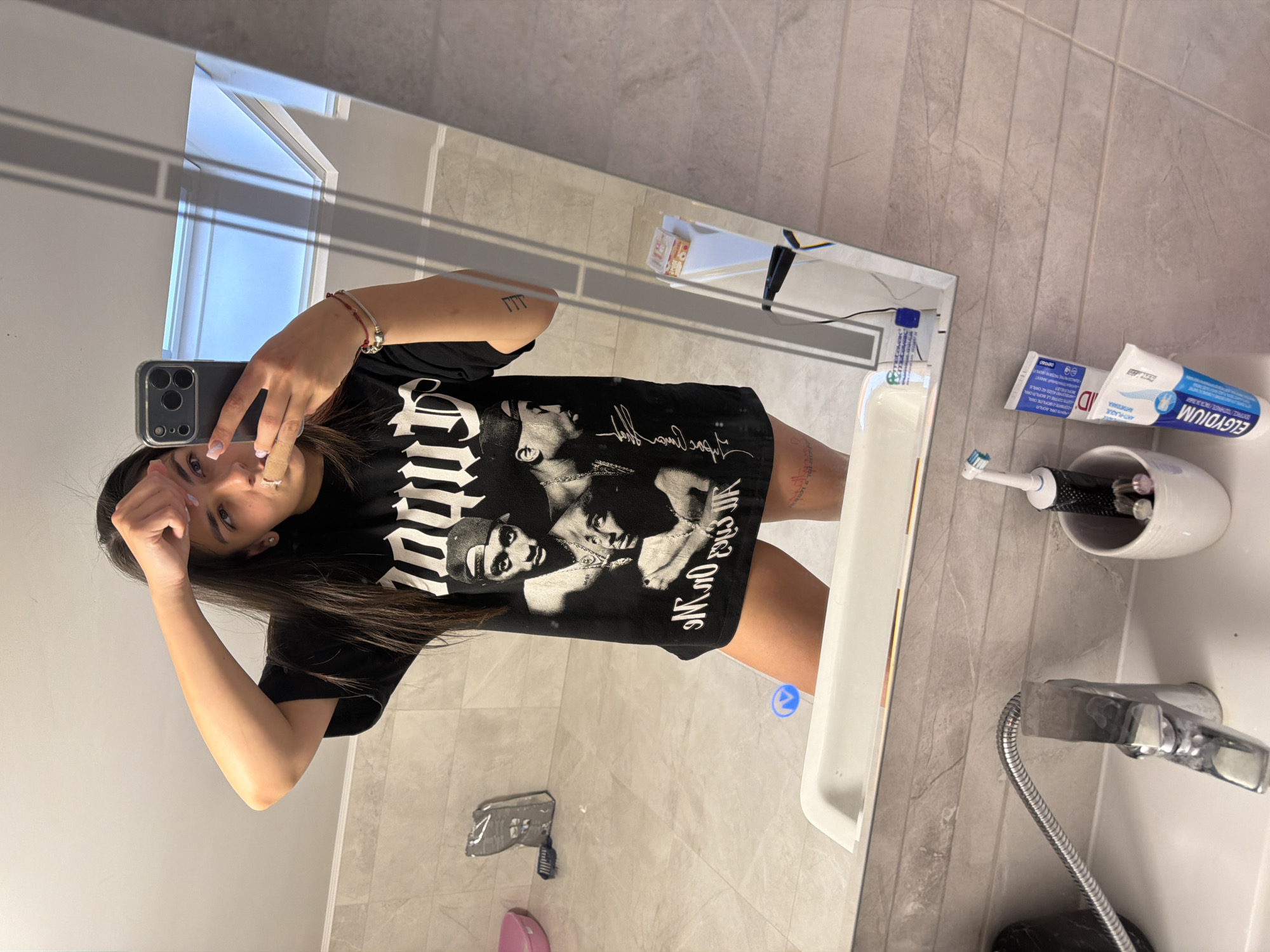 A woman is taking a selfie in a bathroom mirror. She is holding a phone in her left hand. She has long dark hair and is wearing a black T-shirt. Reflected in the mirror are a sink, faucet, toothbrush holder with toothbrushes, a tube of toothpaste, a comb, and a pink object on the floor. The bathroom has tiled walls and flooring. The location in the city of Gabrovo, Bulgaria, cannot be verified.