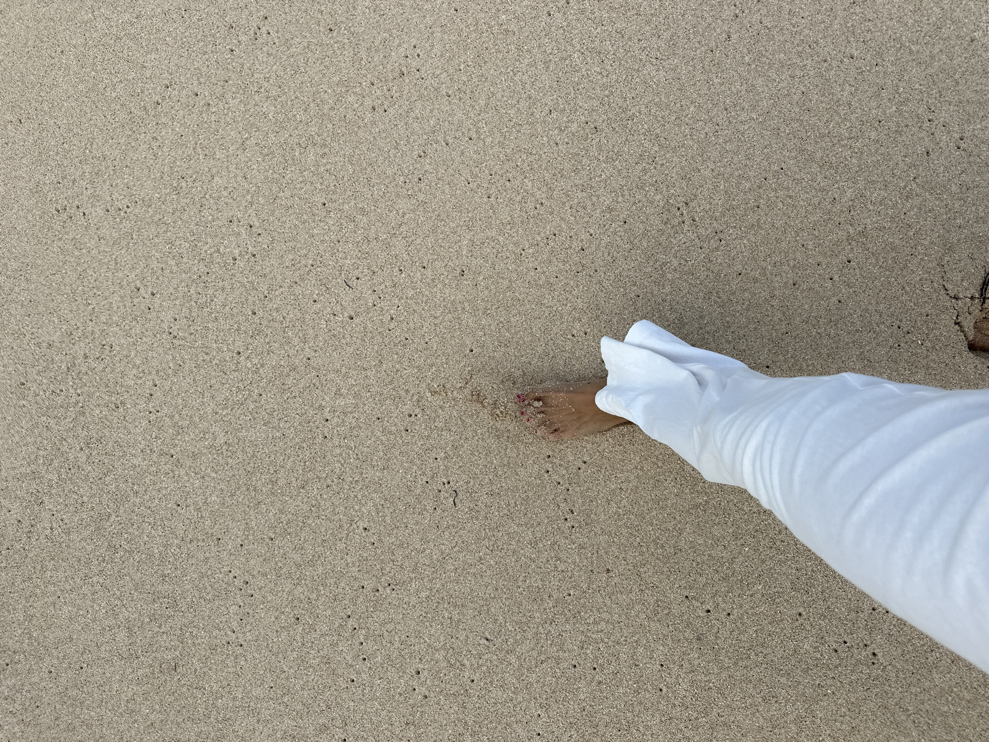 An overhead shot shows a person's left foot and lower leg walking on a sandy beach in Kuta Selatan, Indonesia. The sand is a light brown color and has many small dark holes scattered across it. The person's foot is bare and has painted pink toenails. They are wearing white flowing pants that cover their lower leg. The scene is captured from a high angle, looking down at the sand and the person's leg.