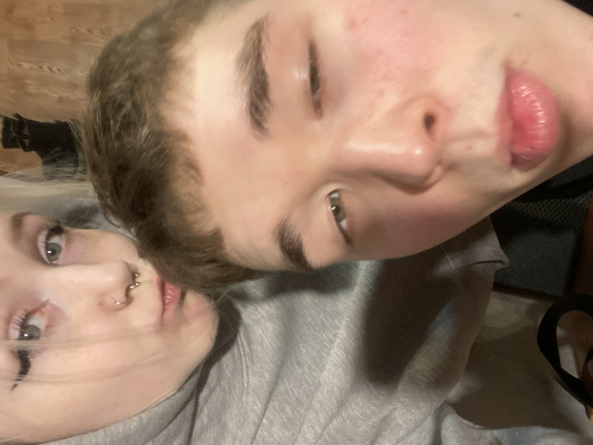 This is a close-up, tilted portrait featuring two individuals, likely young adults, in what appears to be an indoor setting in Izhevsk, Russia.
On the right side of the frame, one person is shown with light brown, slightly wavy hair, fair skin with some visible redness on the forehead and cheeks, and light-colored eyes, one of which is partially closed, conveying a relaxed or sleepy expression. Their nose is prominent, and they have full, pink lips. They are wearing a grey hooded top, partially visible on their shoulder.
The second person, positioned on the left side, has lighter-colored (possibly blonde or light brown) hair that is partially swept away from their face. They have fair skin, light-colored eyes, and a neutral or slightly serious expression. This individual has multiple facial piercings, including a septum ring and a small hoop piercing on their left nostril. A streak of black eyeliner is visible smudged or intentionally applied below their left eye. They are also wearing a grey hooded garment, the fabric of which fills much of the lower frame.
The two individuals are positioned very close to each other, suggesting intimacy or close friendship, possibly in a casual selfie-style shot. The background behind the person on the right features a blurry wooden surface, likely a wall or furniture, indicating an indoor environment. There are no specific time-of-day cues, and no visible text is present in the image.