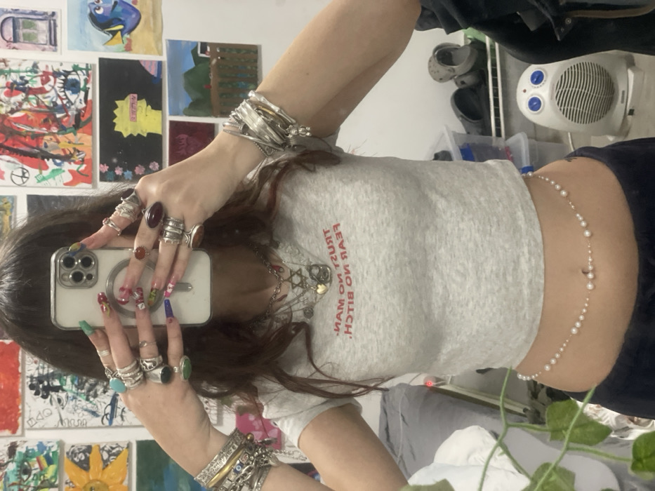 A mirror selfie captures an individual, likely female, within an indoor setting in Raanana, Israel. The person's face is obscured by a white-cased smartphone, held directly in front by both hands. The hands display numerous silver rings, many with large gemstones, and have long, multi-colored manicured nails. Both wrists are adorned with multiple silver bangles and bracelets, including a small bear charm on the right wrist. The individual wears a light grey, short-sleeved crop top with the mirrored red text "TRUST NO MAN FEAR NO BITCH" across the chest. A silver chain necklace with several pendants, including a Star of David and a heart, hangs around the neck. A pearl and bead belly chain featuring a central crescent moon charm is visible around the waist, positioned above dark-colored lower garments. The background wall is white and covered with numerous unframed artworks, depicting varied subjects such as a sunflower, a peace sign, a yellow cartoon character resembling Bart Simpson, and Dory from Finding Nemo. In the lower right portion of the frame, a white electric fan/heater, plastic storage bins, and a dark shoe are visible on the floor. A green plant is partially present in the lower left foreground.