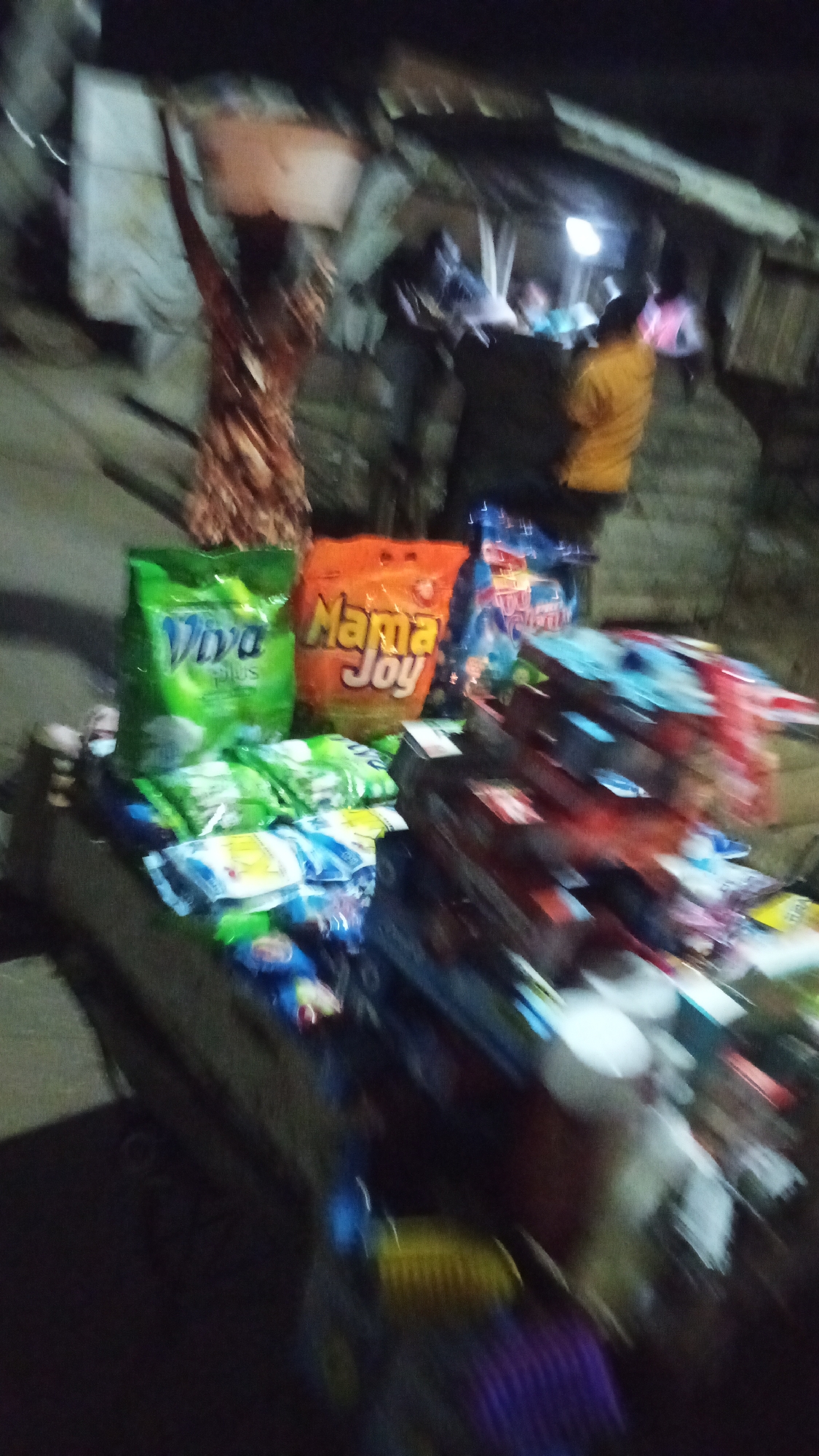 This highly blurred, low-light image depicts a vibrant night scene, characteristic of a street market or informal vending area, likely in Lokoja, Nigeria. The time of day is clearly nighttime, with artificial light sources providing illumination.
In the foreground, a street stall displays various packaged goods. On the left, two large, brightly colored bags are prominent: a green one with "Viva plus" visibly printed on it, and an orange one clearly labeled "Mama Joy." Below these, smaller, individual packets are arranged, possibly containing detergents or snack items. To the right, a stack of what appears to be numerous boxes and packages is visible, though too indistinct due to the blur to identify specific products.
The background is populated by several blurry human figures. On the far left, a person is seen carrying an item on their head, a traditional method of transporting goods. Further back, other individuals are present, including one wearing a yellow shirt and another with a pink top, appearing to move or gather within the market space. A single, bright, unshaded light bulb casts a strong glow in the mid-ground.
The structures in the background appear to be simple, possibly temporary stalls constructed from wood and basic materials, common in informal market settings. The scene conveys an atmosphere of commerce and activity in a lively, albeit dimly lit and bustling, outdoor environment. The pervasive blur throughout the image suggests either significant motion or camera shake during the capture, likely exacerbated by the low light conditions.