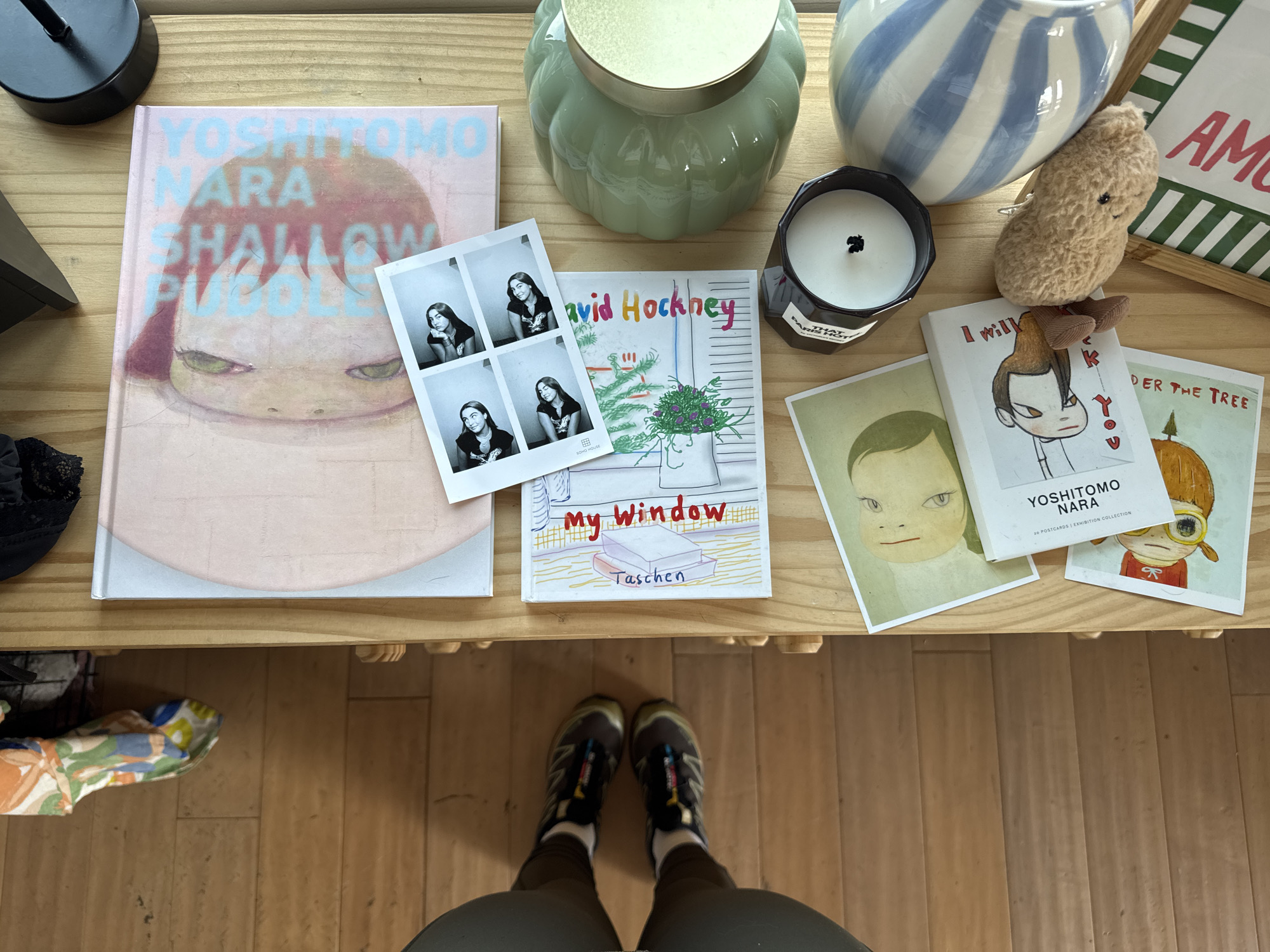 Here is a description of the image:
The image shows a collection of books and decorative items arranged on a light wooden surface, presumably a table. The books feature art by Yoshitomo Nara and David Hockney. A photo booth strip with four images of a person is visible. Other items include a candle, a plush toy, and a decorative vase. The viewpoint is from above, looking down onto the tabletop and beyond to the lower torso and feet of an individual standing in front of the table. The person is wearing dark-colored athletic shoes and olive-green pants.
It is not possible to determine the location as Los Angeles, United States of America based on the contents of the image. The image mainly consists of the objects on the table and a partial view of a person's lower body. There are no specific landmarks, signs, or other visual elements that would help to identify the location of the photo.
