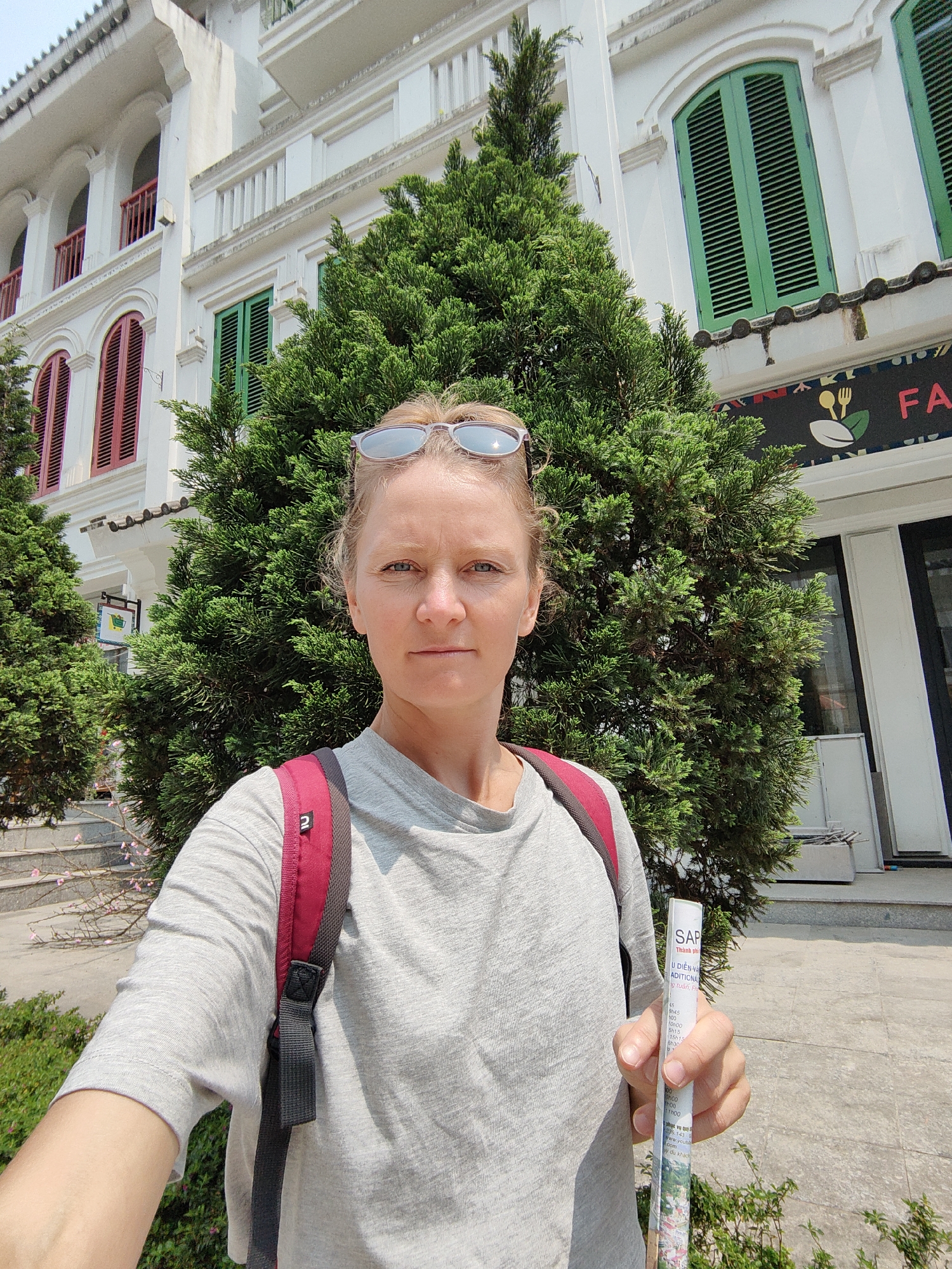 An adult female is depicted in a selfie, facing the camera with a neutral expression. She has light-colored hair, with sunglasses positioned on top of her head. She wears a light grey short-sleeved t-shirt and a red and black backpack, with one strap visible over her right shoulder. In her extended right hand, she holds a rolled-up white leaflet, partially visible with the word "SAPA" and Vietnamese text.
The background features a multi-story white building with distinct architectural elements, indicating the location is in the city of Sa Pa, Vietnam. The building exhibits arched windows with green louvered shutters on the right side and red louvered shutters on the left side. Balconies with white and red railings are also visible. A large, dense green conifer tree stands directly behind the woman, partially obscuring the building's facade. A smaller conifer tree is visible to the left, alongside steps and sparse pink blossoms. A sign with a spoon, fork, and leaf icon, along with the partial letters "FA", is visible on the building's ground floor to the right of the main tree. Another sign with a shopping cart icon is visible further left. The scene is illuminated by bright daylight.