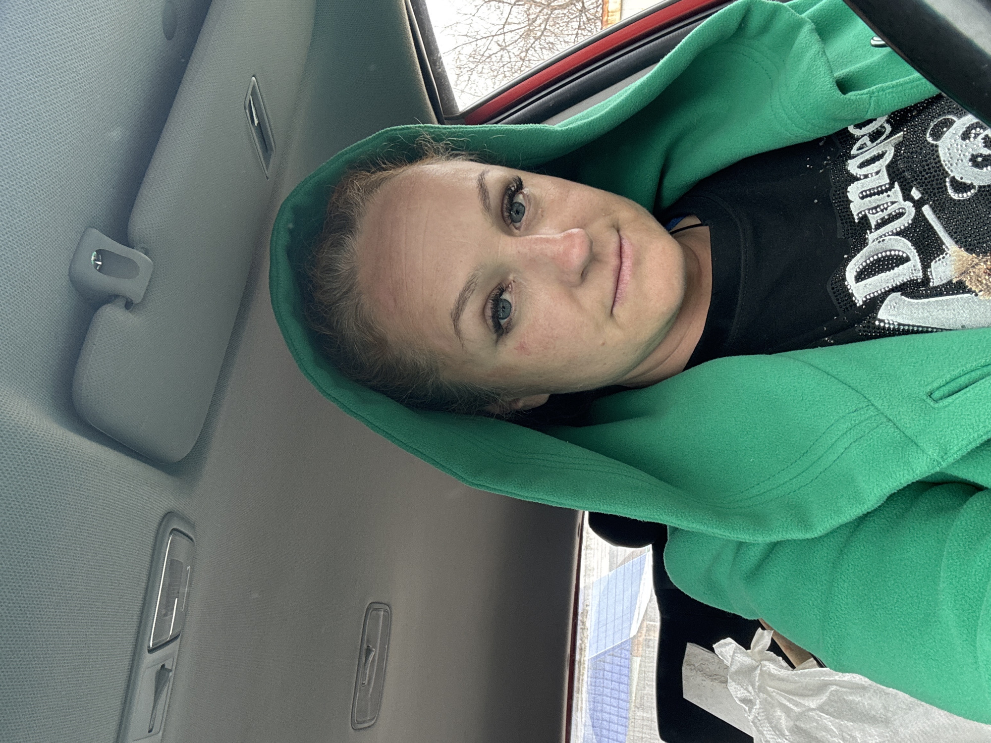 A woman is inside a car. She wears a green hooded jacket over a black shirt. The interior of the car is grey. Outside, the branches of a tree are visible. The woman appears to be in Chaikovsky, Russia.