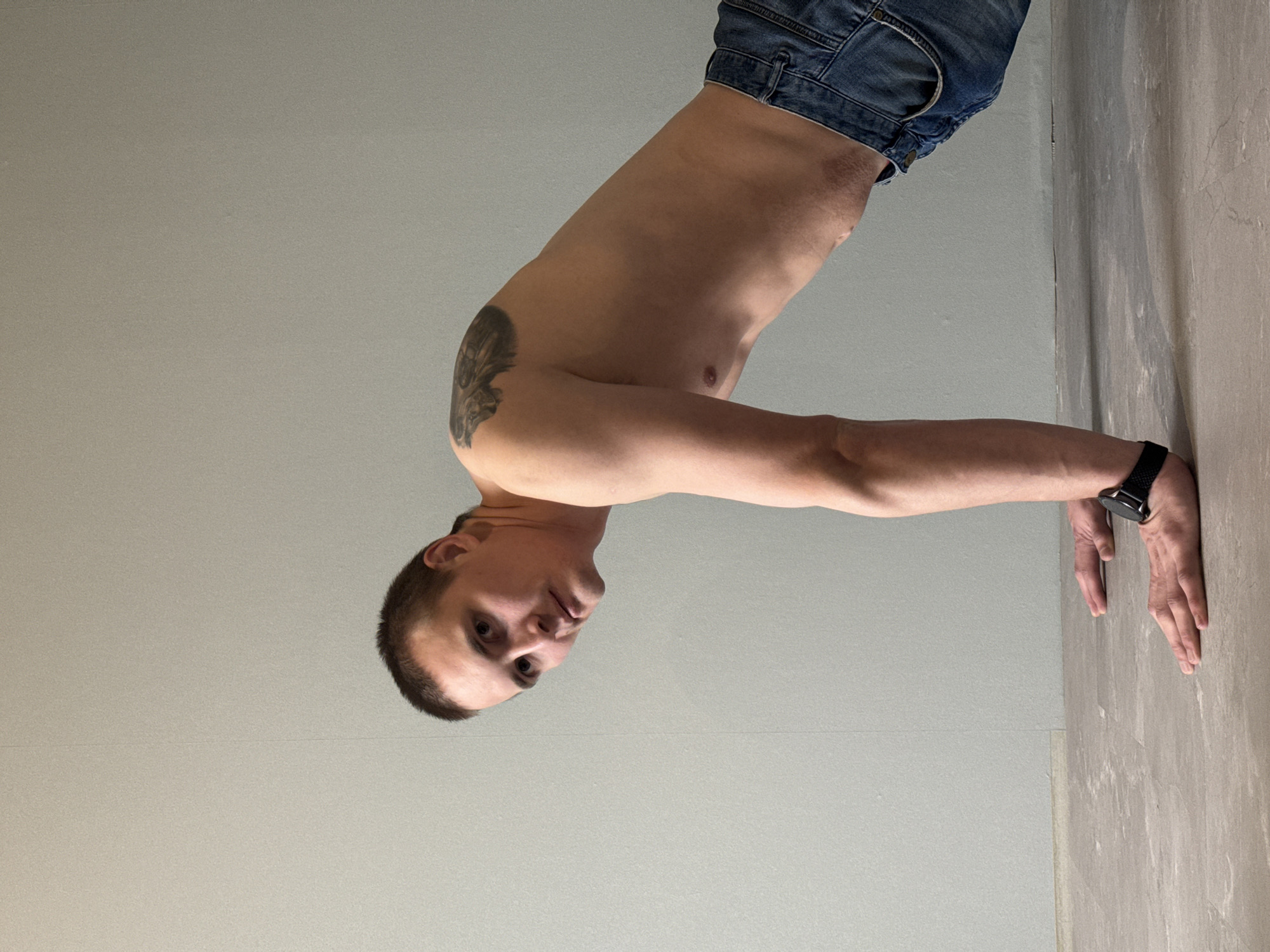 This media file captures a man performing an inverted physical exercise, such as a handstand or a similar bodyweight movement, indoors in Chaikovsky, Russia. The man, who appears to be young to middle-aged with short, dark hair, is shown from the waist up. He is shirtless, revealing a toned torso and a prominent tattoo on his left upper arm/shoulder. He is wearing blue denim jeans and a dark-colored smartwatch on his left wrist.
His hands are placed firmly on a textured, light brown-grey surface, which forms a corner with a plain, light grey wall. He is looking directly at the camera with a neutral expression. The setting is minimalist, with simple, unadorned walls, suggesting a functional space like a gym, studio, or a room under construction/renovation. The lighting is even, typical of indoor conditions, providing no specific indication of the time of day or outdoor weather. There is no visible text in the image.