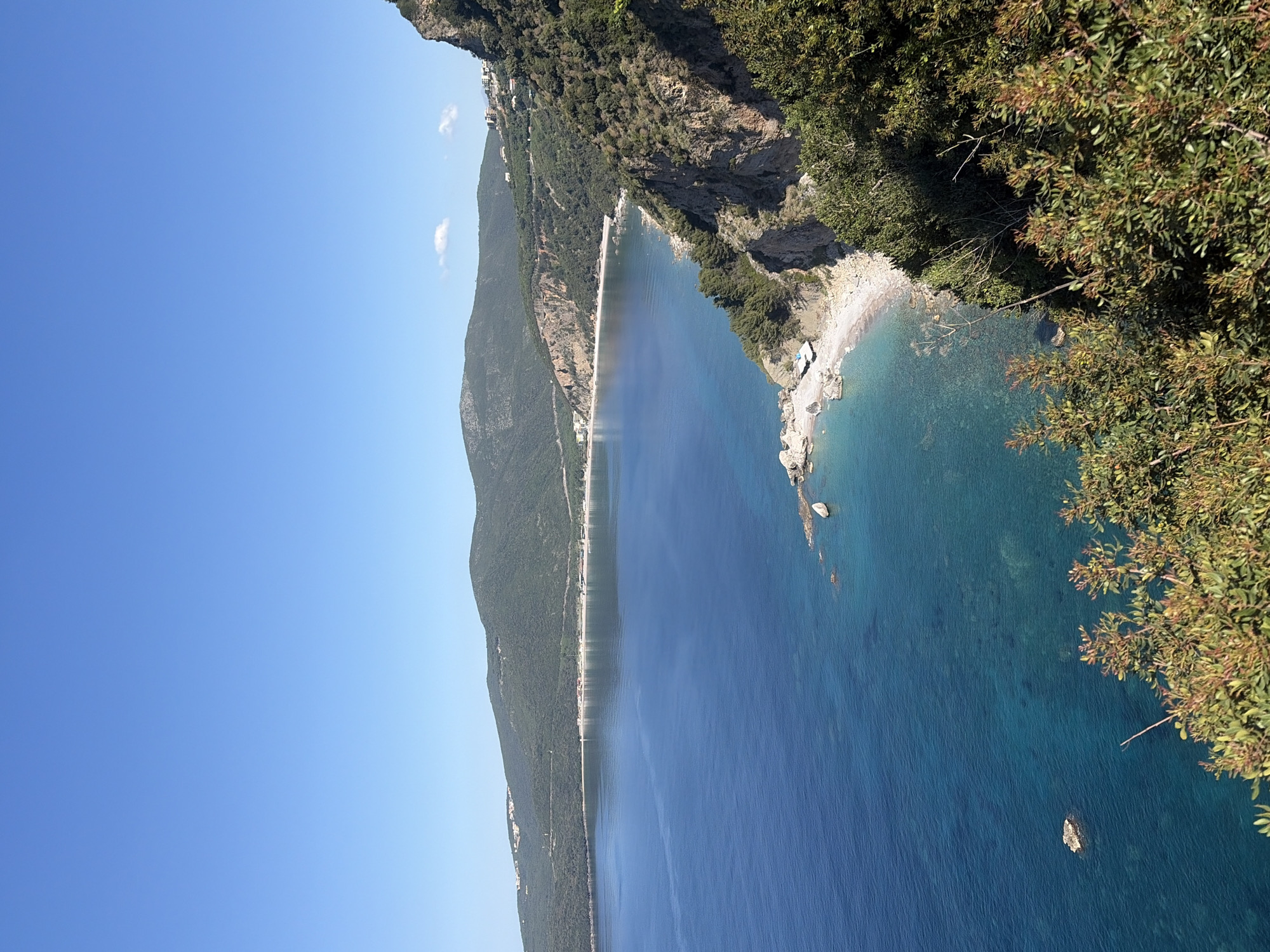 A scenic view captures the coastline near Budva, Montenegro, featuring a rugged cliff descending to the Adriatic Sea. The water transitions from a lighter turquoise near the shore to a deep blue in the distance. A stretch of beach borders the water, with a road and hillside vegetation paralleling the coastline. A clear blue sky dominates the upper left portion of the image. Vegetation clings to the cliff edge in the foreground.